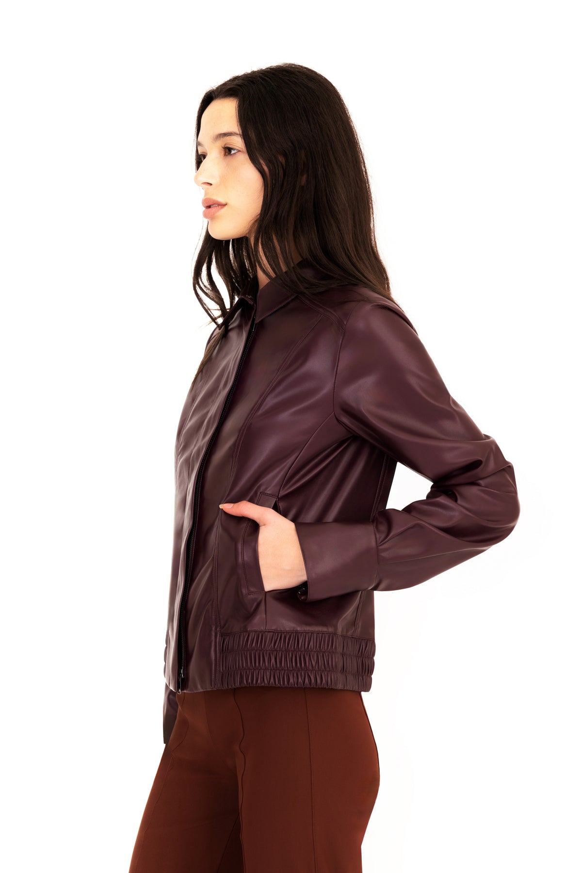 A woman modeling a burgundy vegan leather jacket with a collar, front zipper, and side pockets, paired with brown pants, against a plain white studio background.