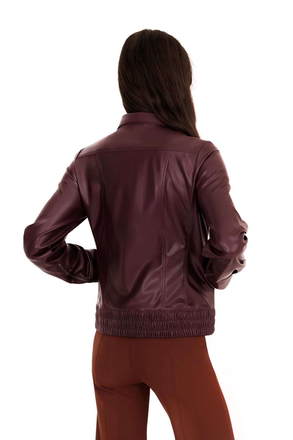 A woman viewed from the back wearing a burgundy vegan leather jacket with a gathered elastic hem, paired with brown pants, against a plain white background.