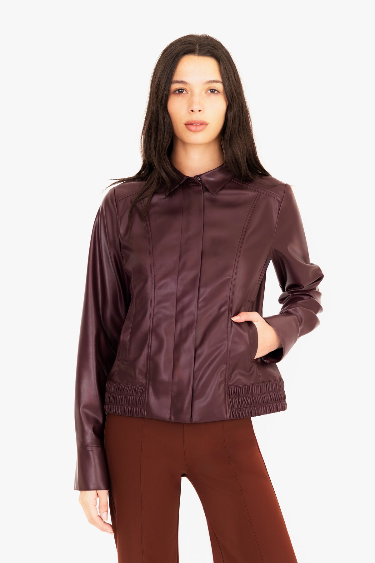A woman modeling a burgundy vegan leather jacket with a collar, front zipper, and side pockets, paired with brown pants, against a plain white studio background.