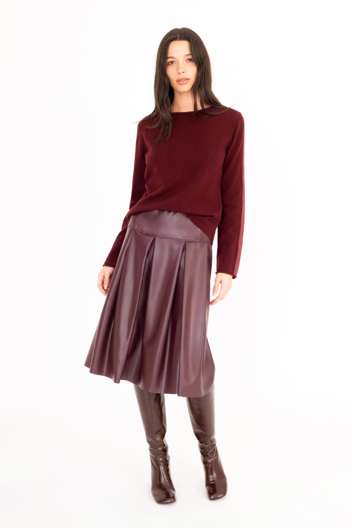 A woman modeling a dark burgundy cashmere knit long-sleeve top and a pleated dark burgundy vegan leather midi skirt, paired with dark brown knee-high boots, against a solid white studio background.