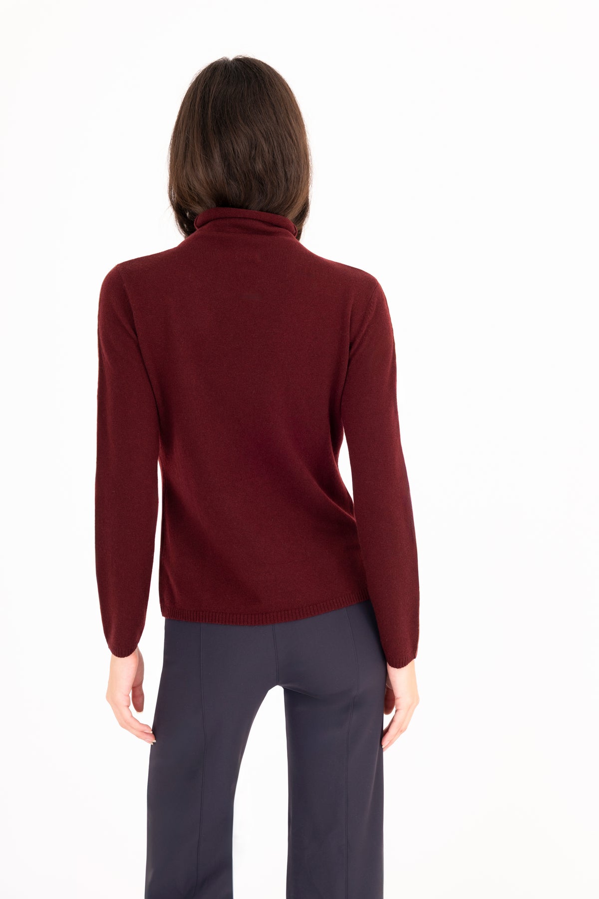 A woman viewed from the back wearing a dark burgundy mock neck sweater and dark navy trousers, set against a plain white studio background.