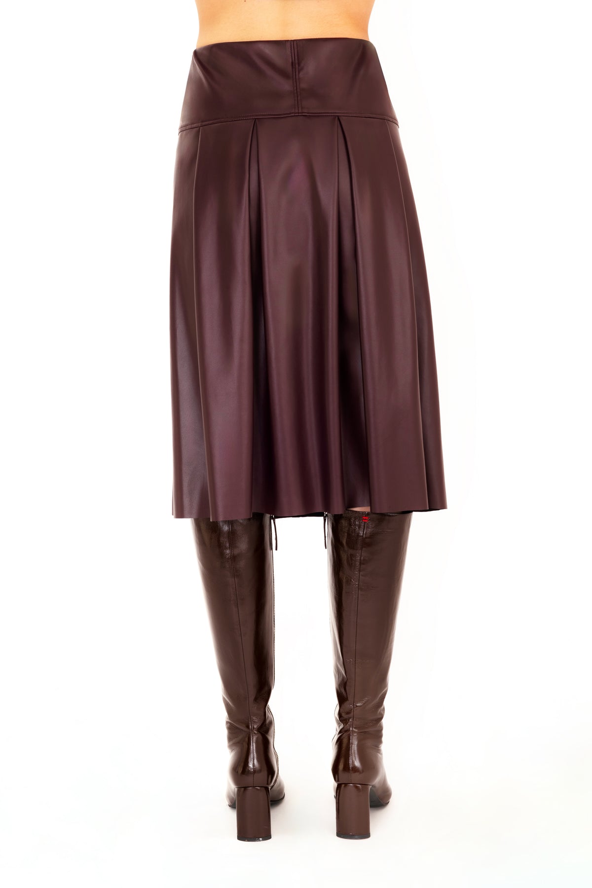Back view of a burgundy vegan leather pleated A-line skirt shown on a model from the waist down, paired with dark brown knee-high leather boots against a plain white background.