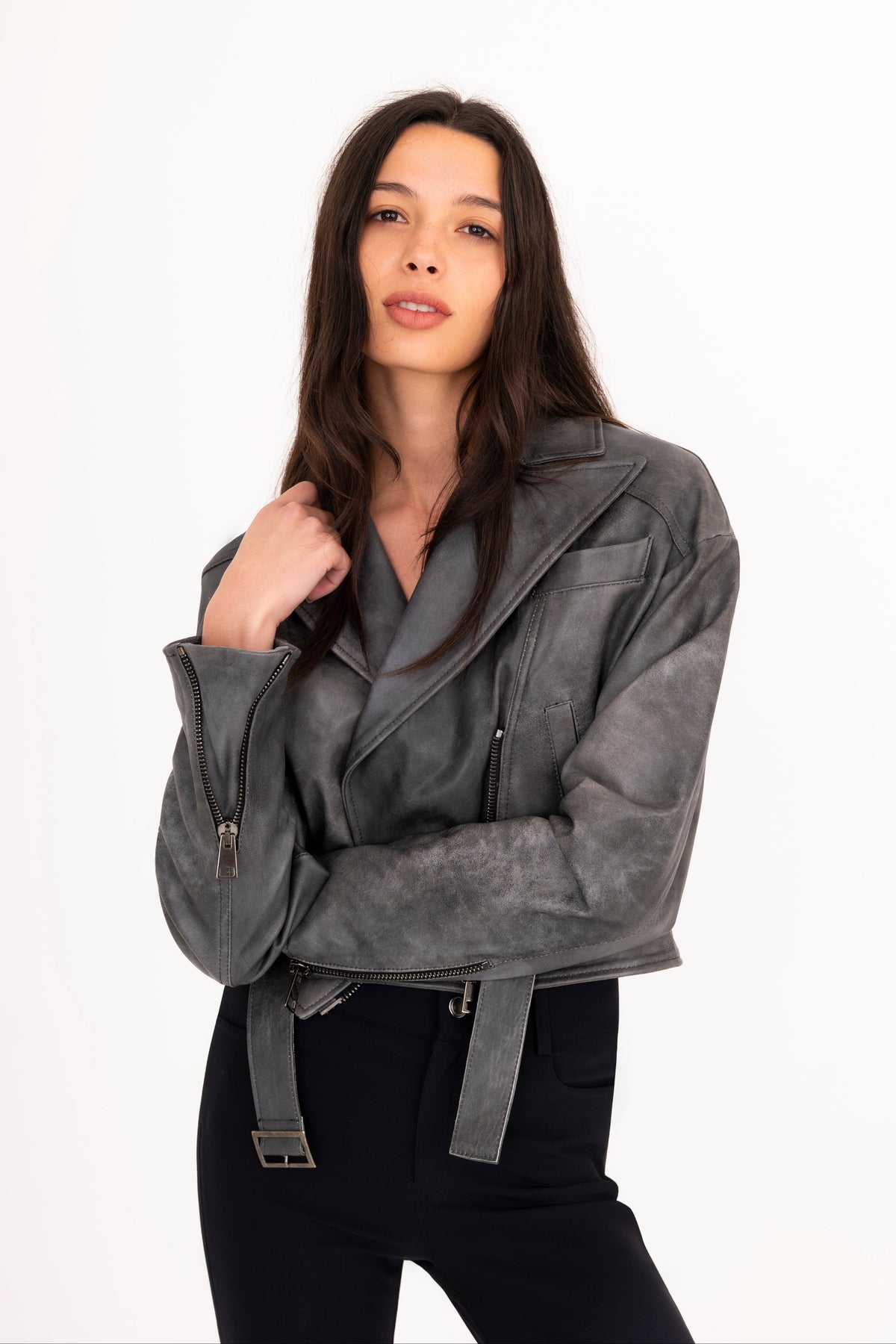 A woman modeling a gray rustic leather moto jacket with a belted waist, paired with black pants, against a plain white studio background.