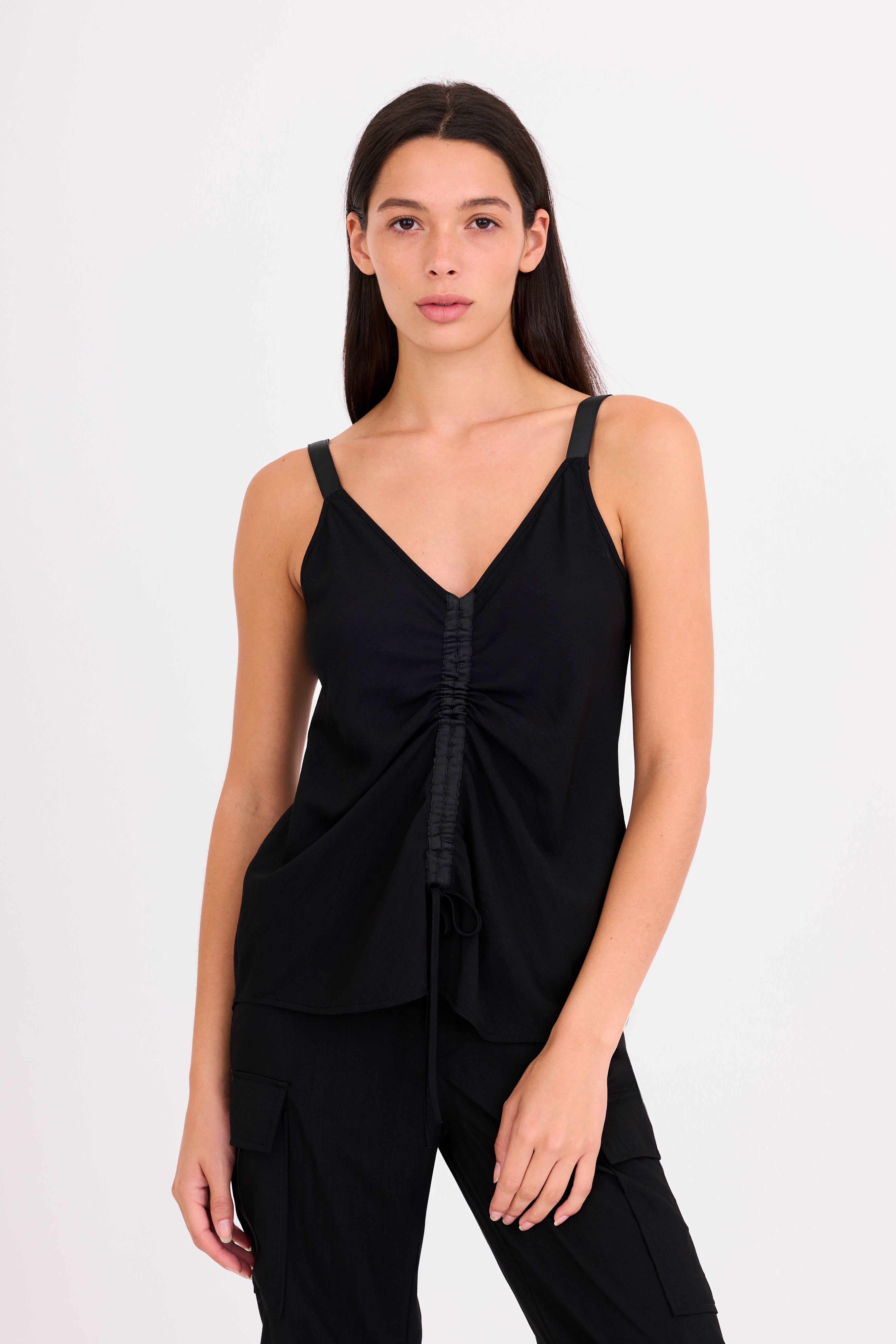 A woman modeling a black sleeveless V-neck top with gathered front detailing and black pants against a plain white studio background.