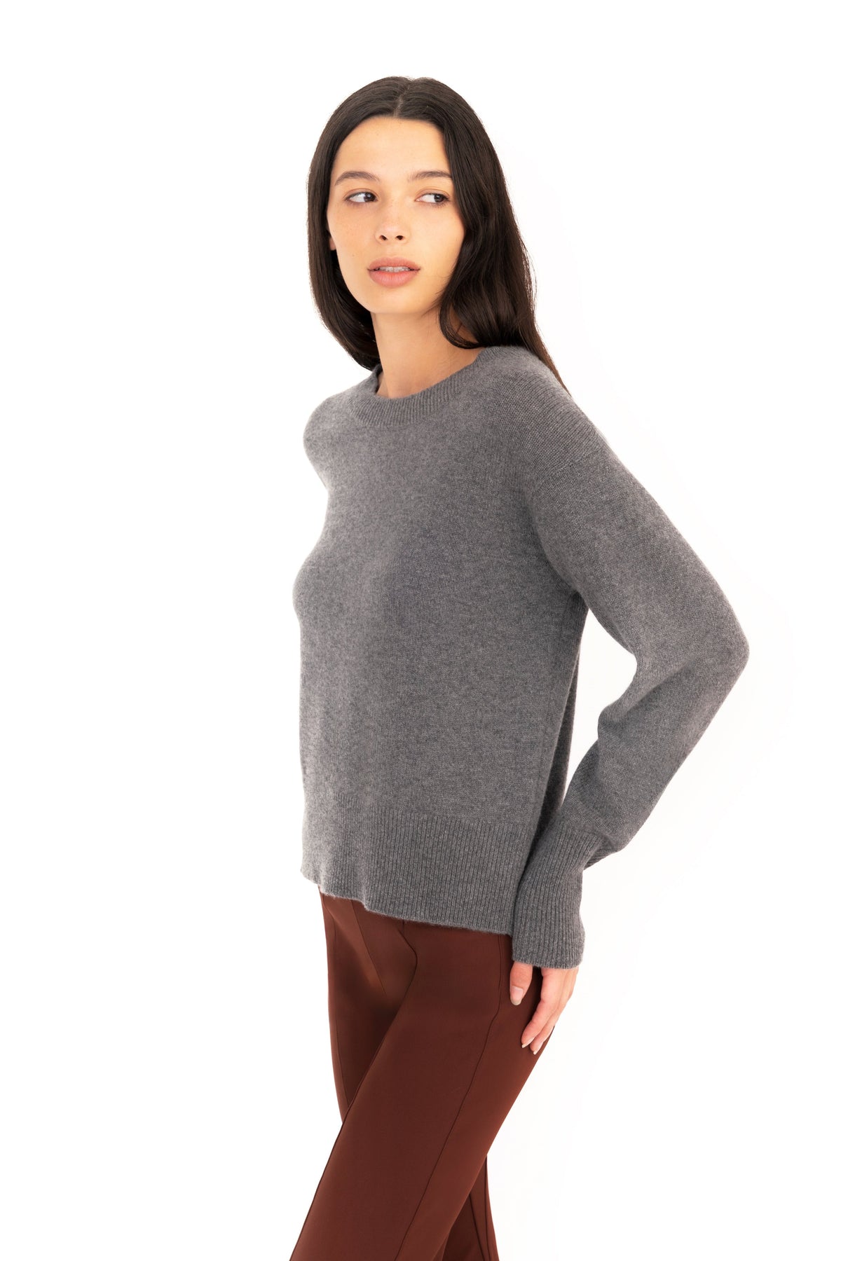 Woman wearing a gray crew-neck sweater and dark brown pants on a white studio background.