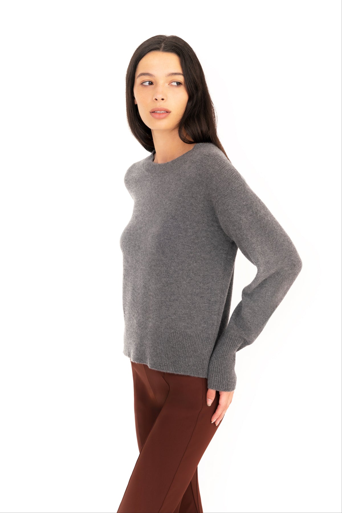 Woman wearing a gray crew-neck sweater and dark brown pants on a white studio background.