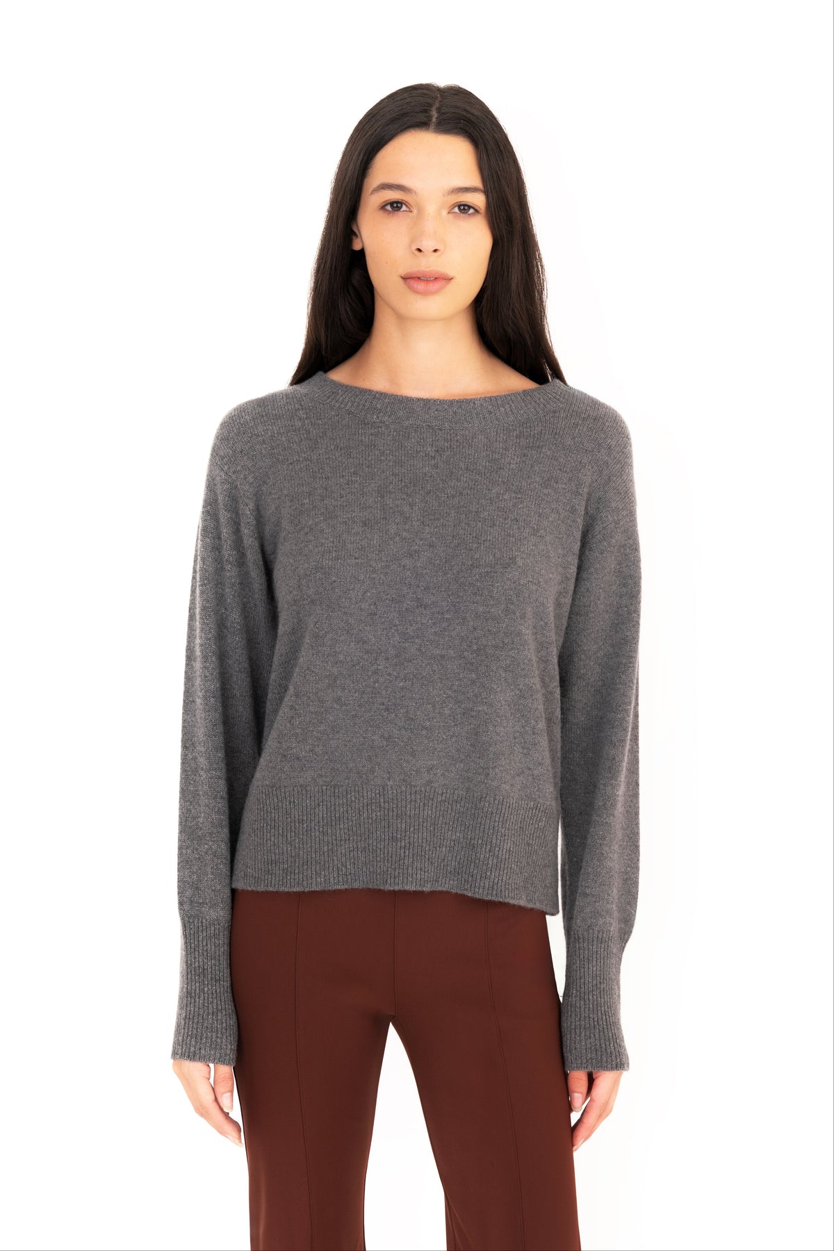 A woman modeling a gray knit crew-neck sweater, paired with dark brown pants, against a plain white background.