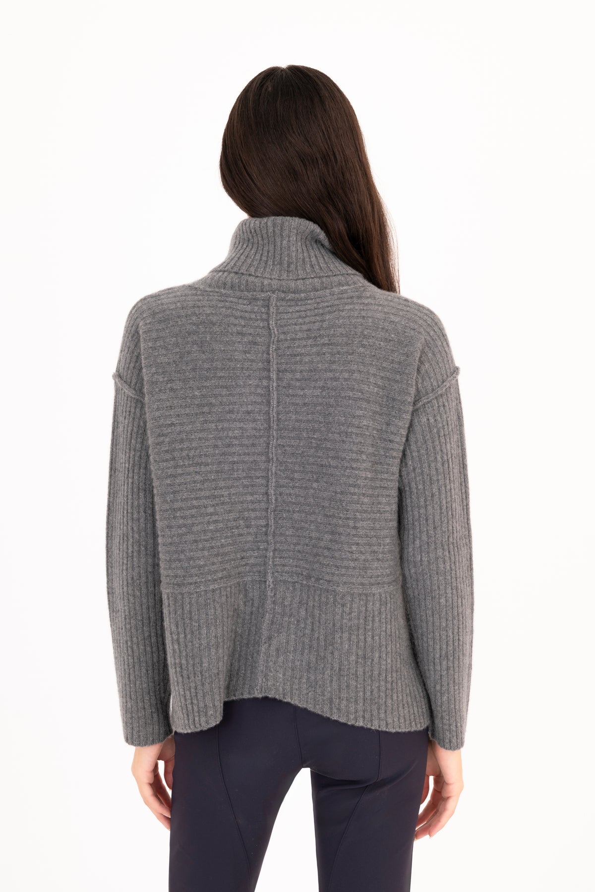 A woman viewed from the back wearing a gray ribbed knit funnel-neck sweater and dark navy pants against a plain white studio background.
