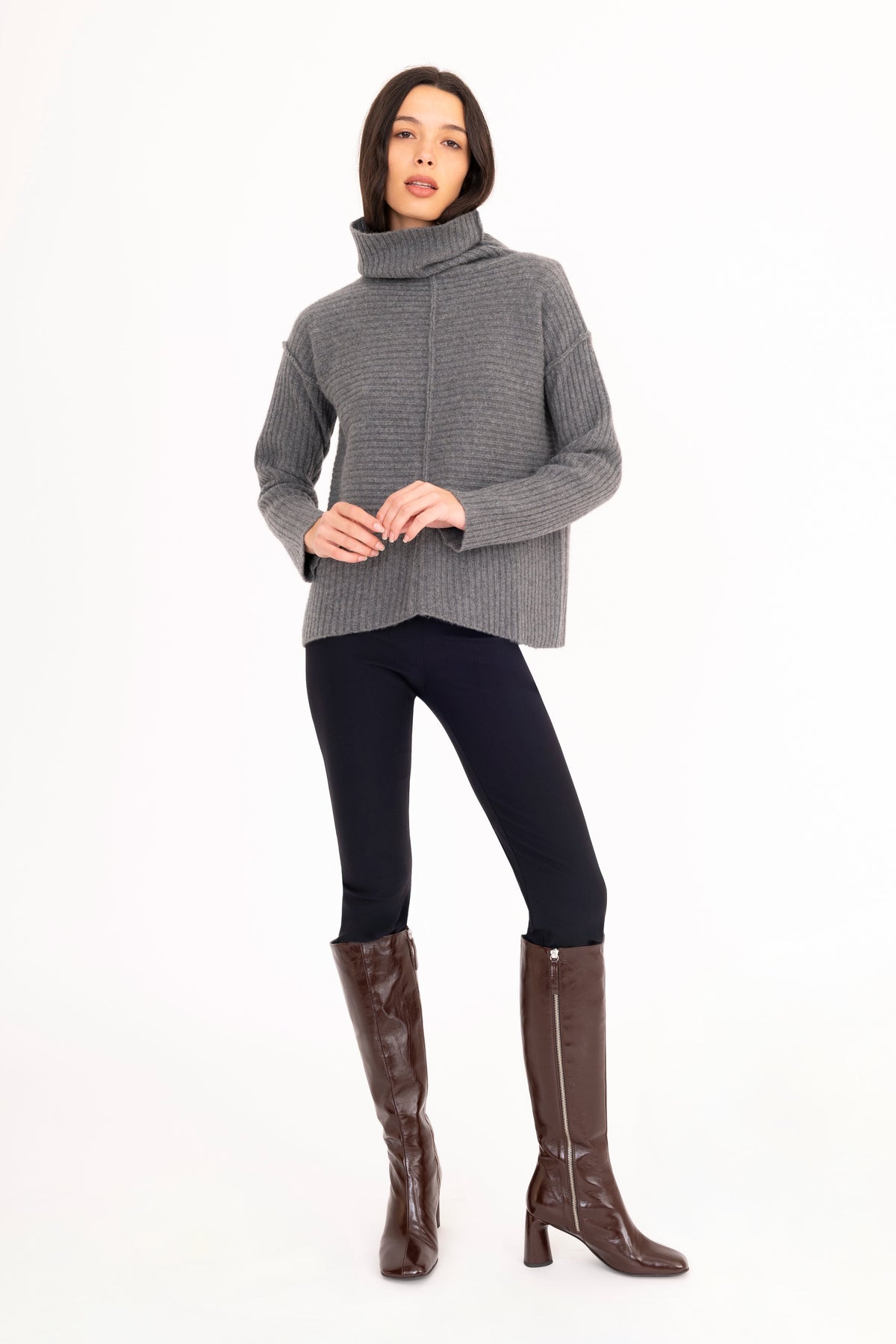 A woman modeling a gray cashmere ribbed turtleneck sweater and black pants against a plain white studio background.