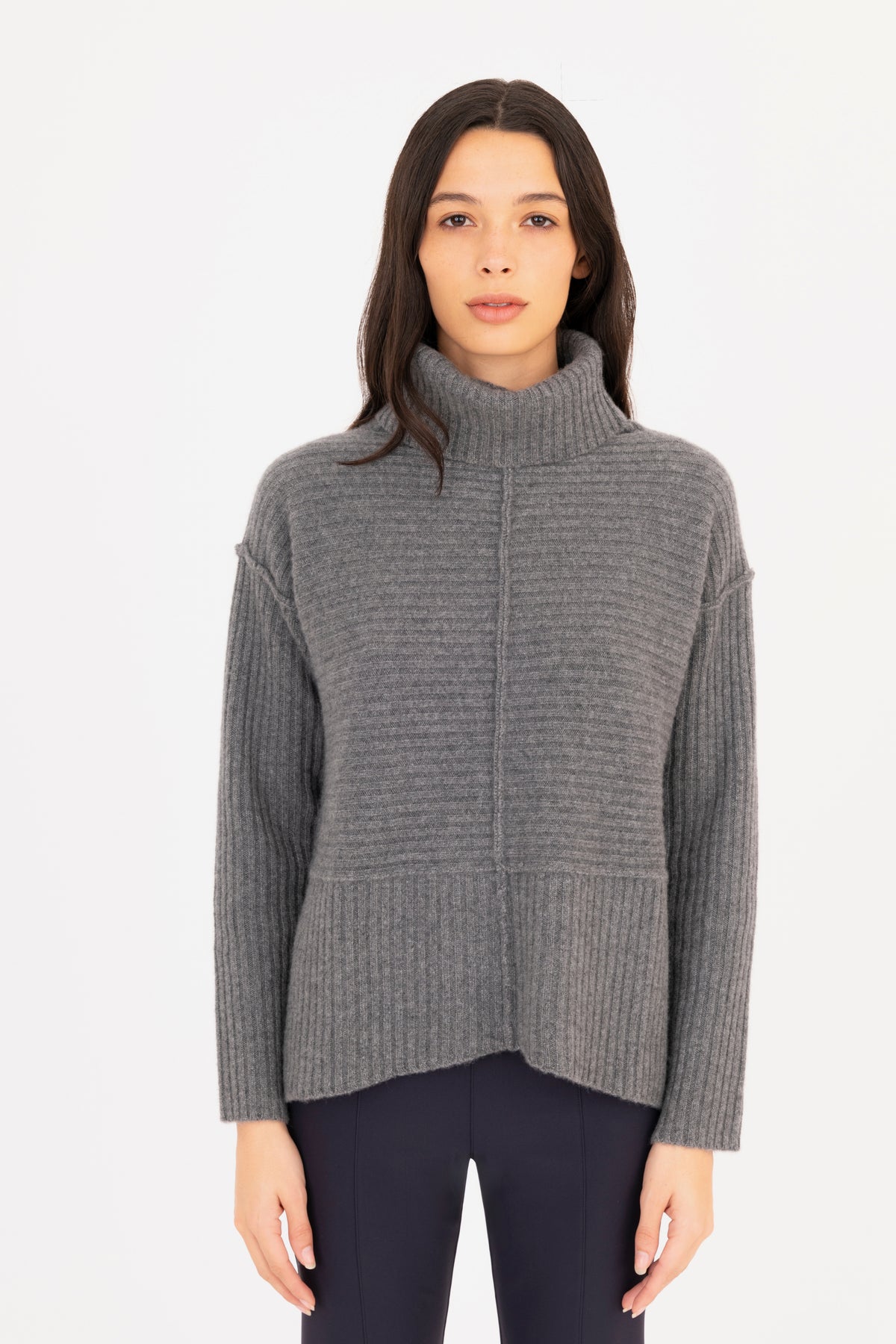 A woman modeling a gray ribbed knit funnel-neck sweater, paired with dark navy pants against a plain white studio background.
