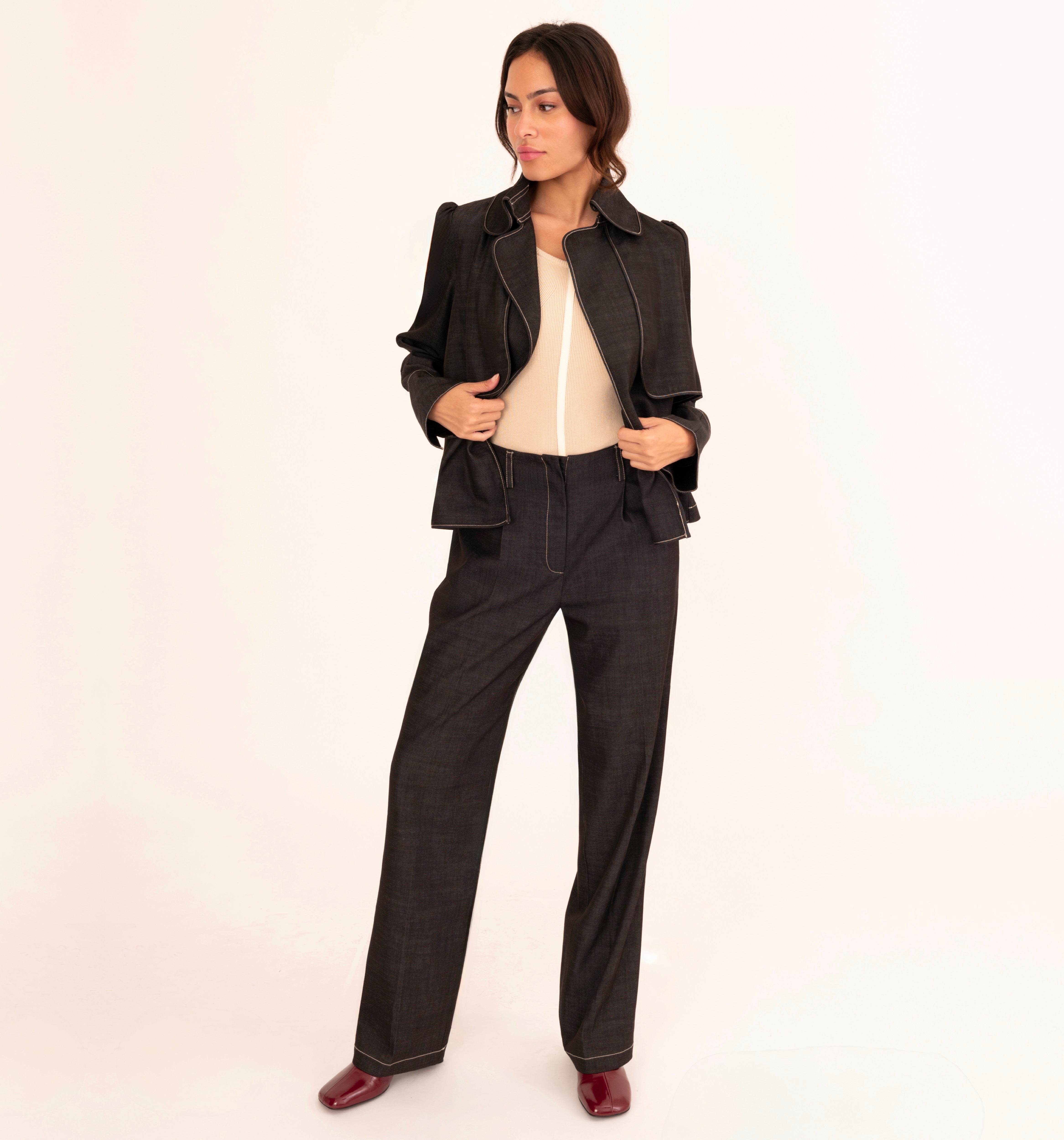 A woman modeling a black two-piece suit consisting of a cropped jacket with structured shoulders and matching wide-leg trousers, styled over a cream top and burgundy shoes, against a plain off-white studio background.