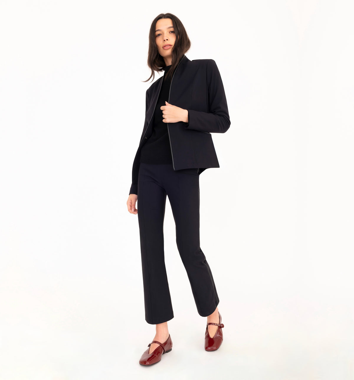 A model wearing a black tailored pantsuit with a cropped blazer, black top, and black cropped trousers, styled with dark burgundy Mary Jane loafers, against a solid white studio background.