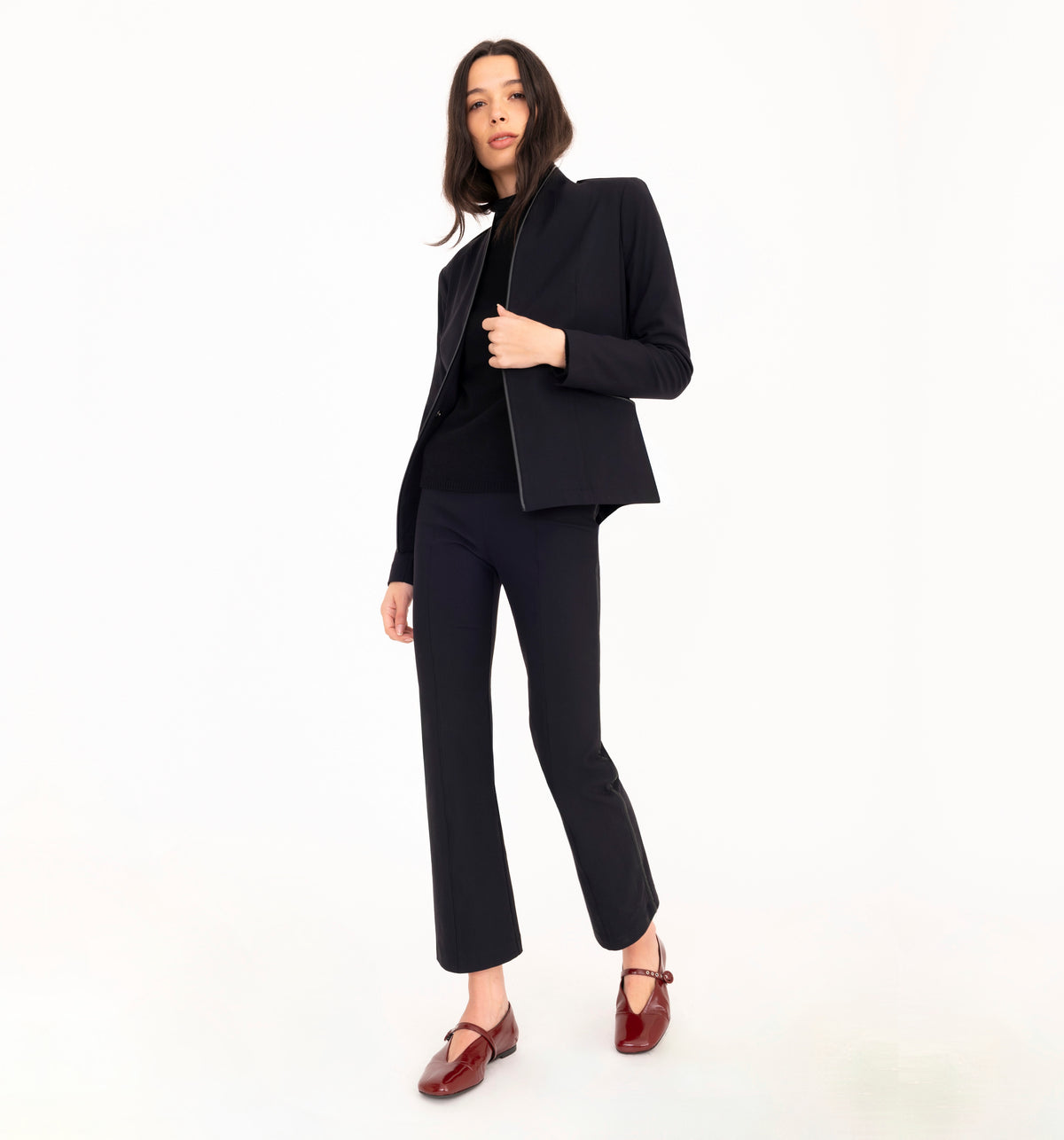 A model wearing a black tailored pantsuit with a cropped blazer, black top, and black cropped trousers, styled with dark burgundy Mary Jane loafers, against a solid white studio background.