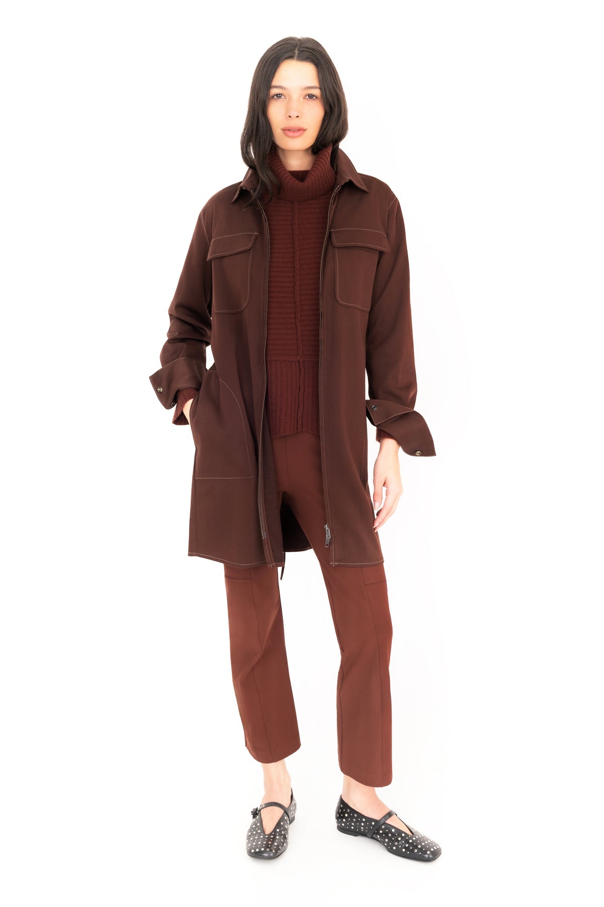 A model stands against a solid white background wearing a matching dark brown three-piece clothing set consisting of a long-sleeve button-up jacket, a ribbed turtleneck sweater, and cropped trousers, paired with black patterned flat shoes.