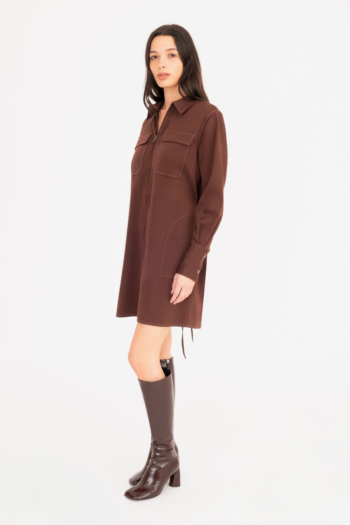 A woman modeling a brown denim shirt dress with front pockets, paired with dark brown knee-high leather boots, against a plain white studio background.