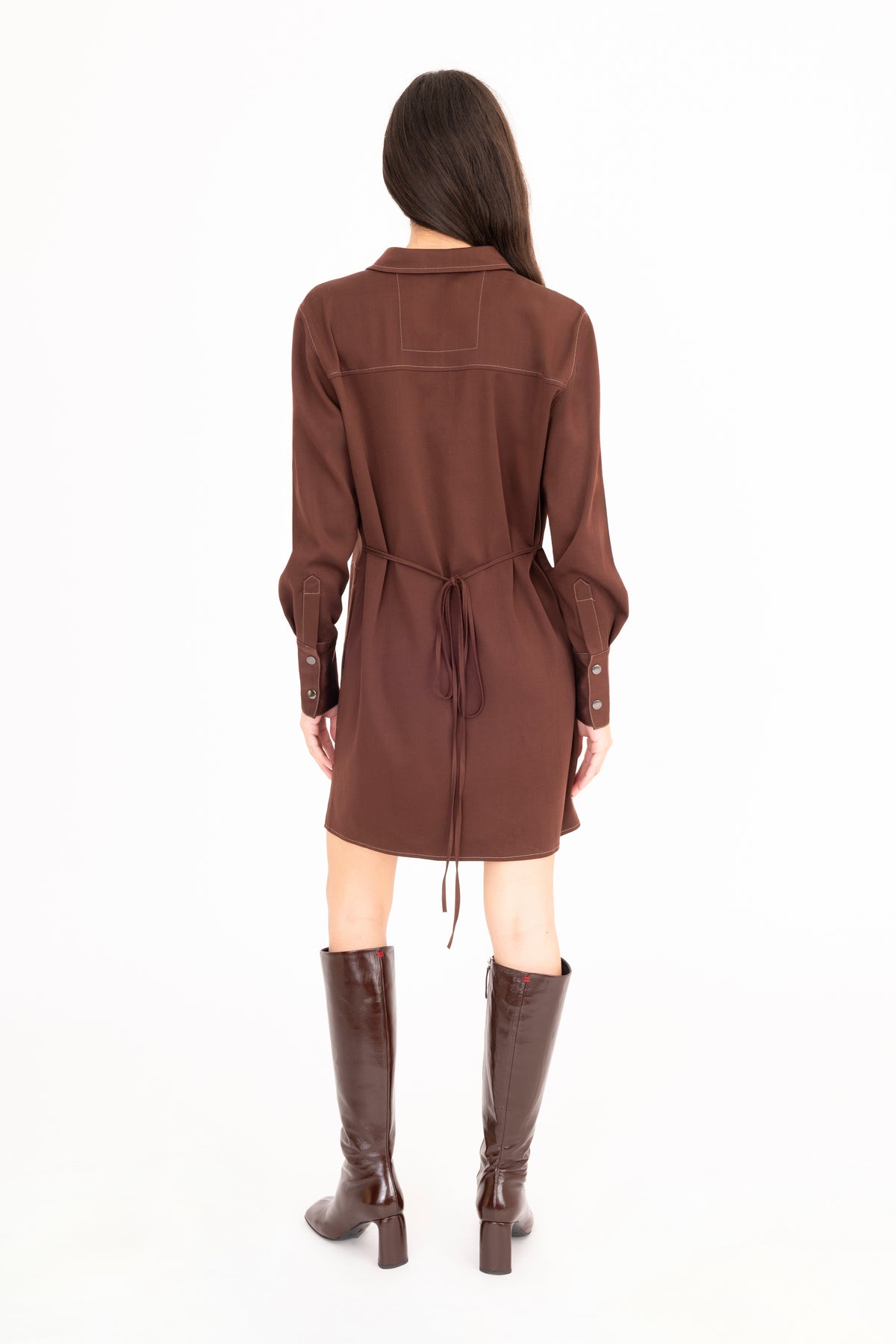 A woman viewed from the back wearing a brown denim belted shirt dress and brown knee-high leather boots, standing against a plain white studio background.