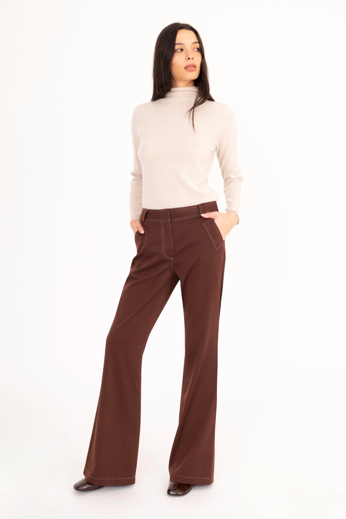 A woman modeling a brown flared denim trousers, and a beige cashmere mock-neck sweater, paired with dark brown leather boots, against a plain white studio background.