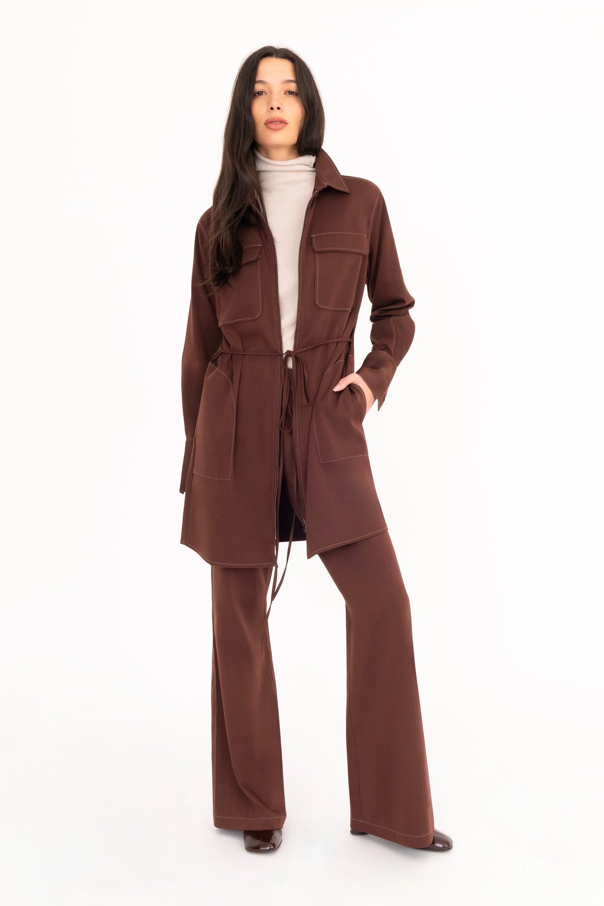 A woman modeling a brown denim shirt dress worn as a jacket with matching flared denim trousers, and a beige cashmere mock-neck sweater, paired with dark brown leather boots, against a plain white studio background.