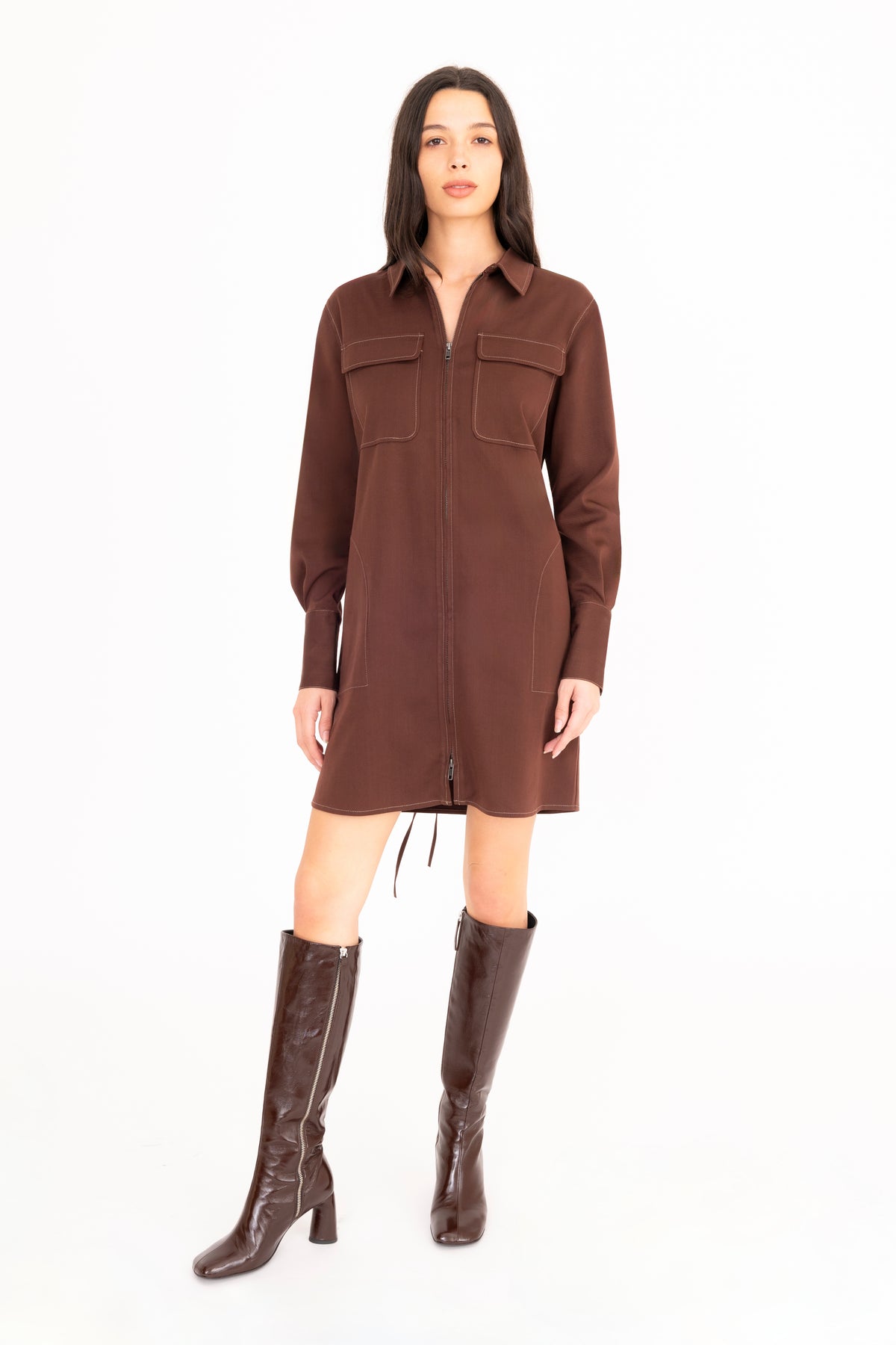 A woman modeling a brown denim shirt dress with front pockets, paired with dark brown knee-high leather boots, against a plain white studio background.