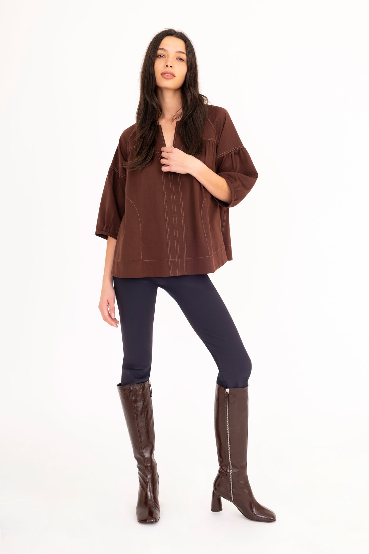 A woman modeling a brown 3/4-sleeve top with vertical seam details, paired with dark navy pants and brown knee-high boots, against a plain white studio background.