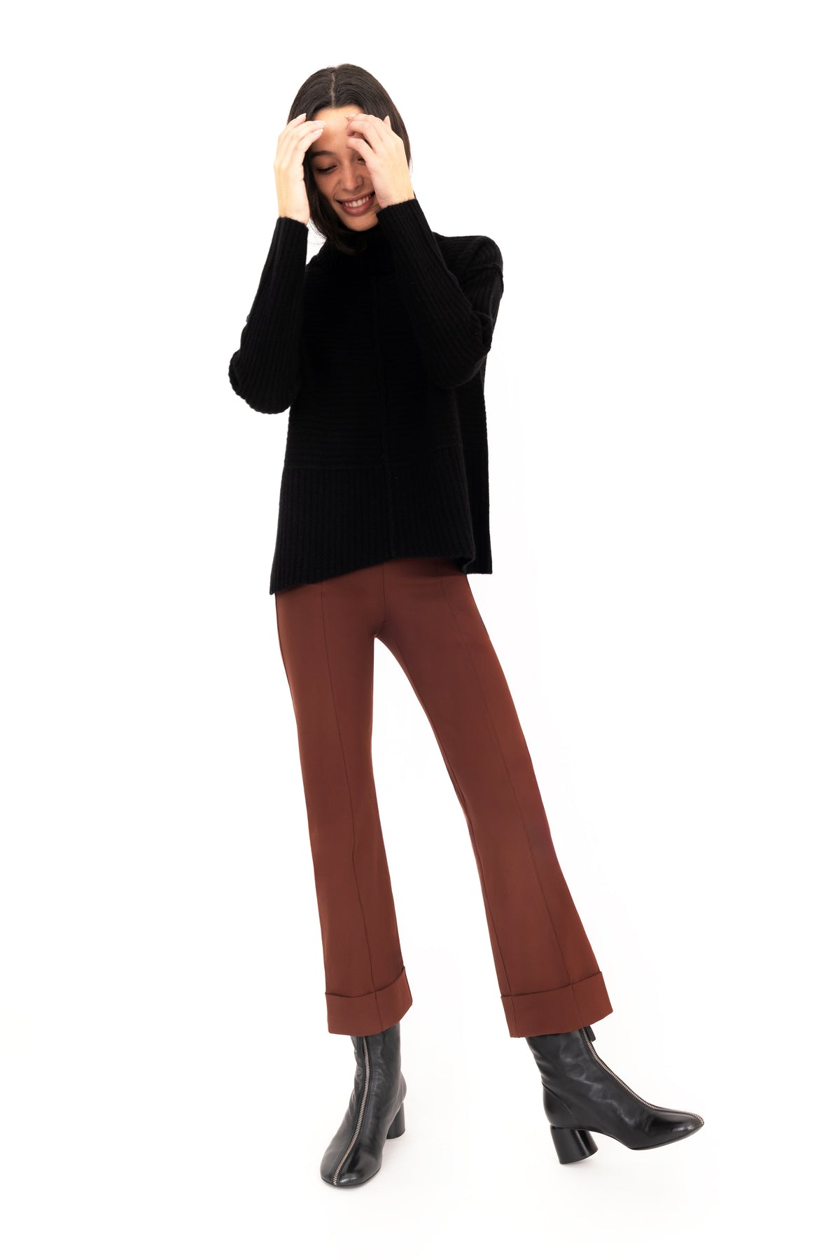 A woman wearing a black cashmere funnel-neck sweater and brown cropped pants on a white background.