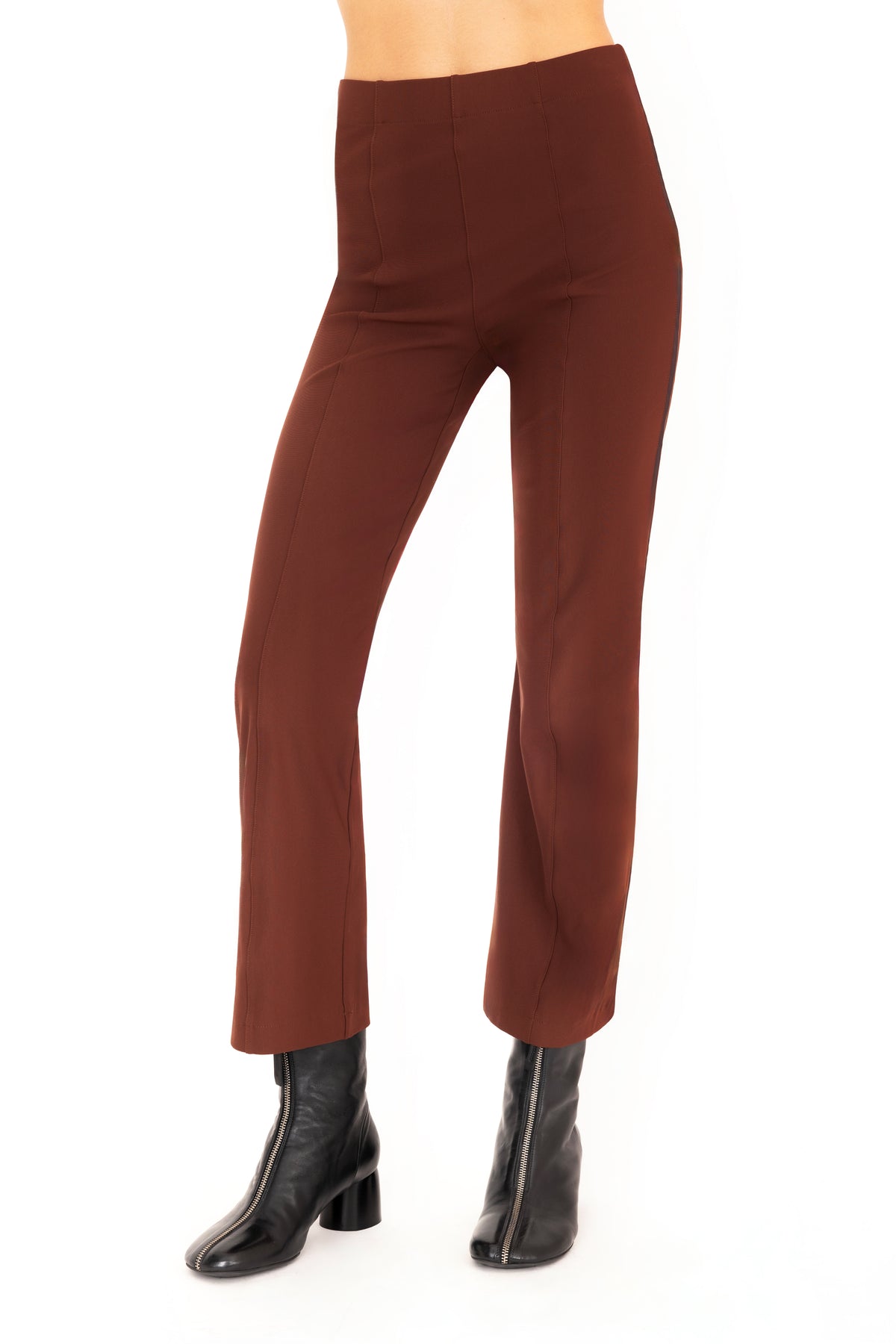 A front view of a model wearing dark brown flared pants with dark brown vegan leather contrast piping and black leather ankle boots, set against a plain white background.