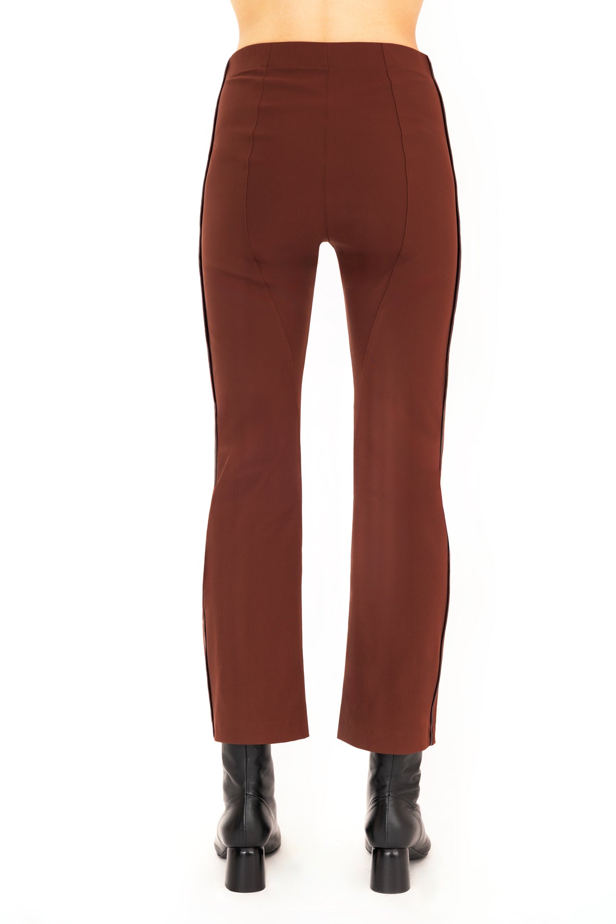 A back view of a model wearing dark brown flared pants with dark brown vegan leather contrast piping and black leather ankle boots, set against a plain white background.