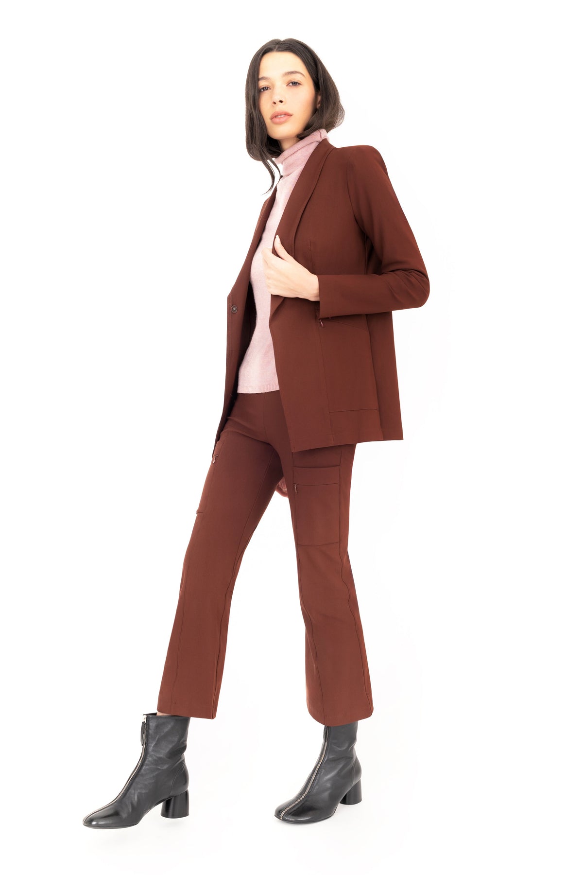 A model wearing a brown tailored pantsuit with a blazer and matching cropped pants, layered over a light pink cashmere turtleneck and paired with black ankle boots, shot against a solid white background.