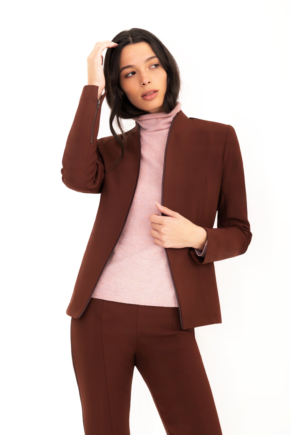 A woman modeling a brown tailored blazer and matching trousers with a light pink cashmere turtleneck sweater, posed against a solid white background.