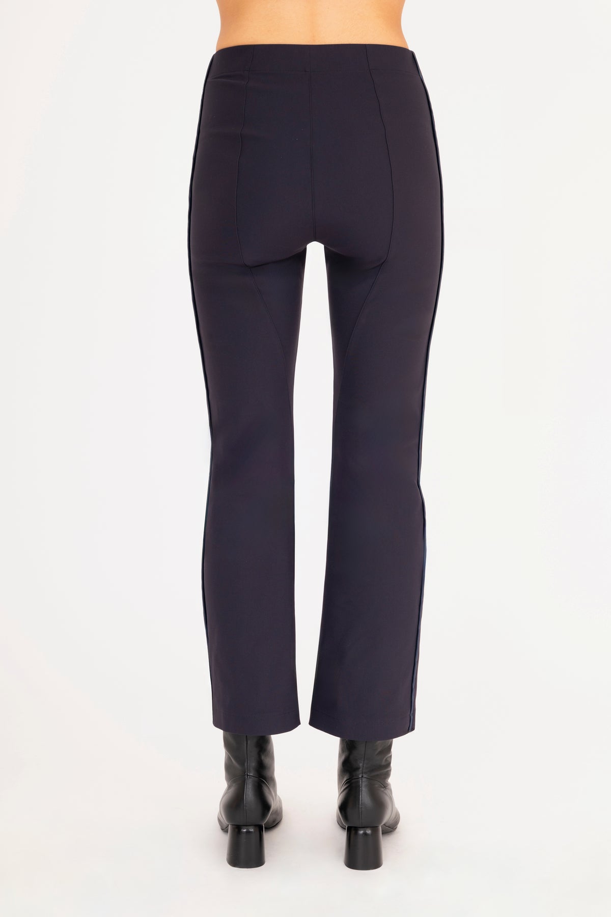 A back view of a model wearing dark blue flared pants with dark blue vegan leather contrast piping and black leather ankle boots, set against a plain white background.
