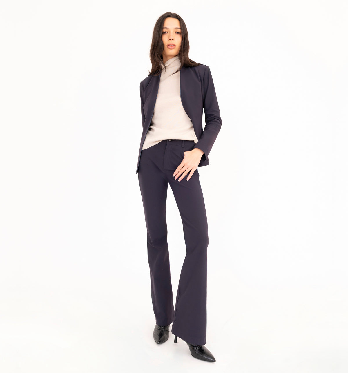 A woman modeling a dark navy blue pantsuit, consisting of a tailored blazer and matching flare jean pants, styled with a beige cashmere mock neck top and black pointed-toe heels against a solid white studio background.