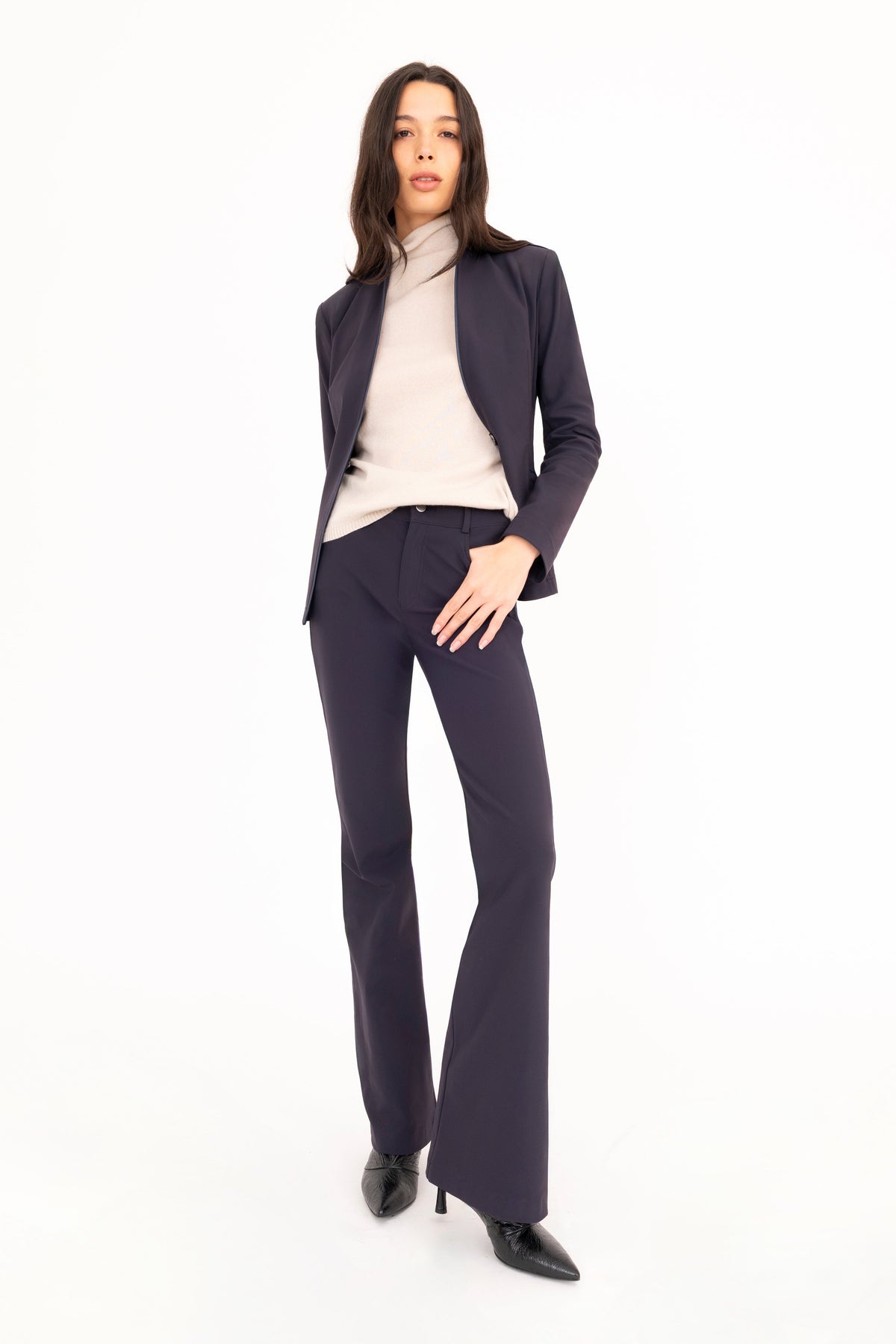 A woman modeling a dark navy blue pantsuit, consisting of a tailored blazer and matching flare jean pants, styled with a beige cashmere mock neck top and black pointed-toe heels against a solid white studio background.