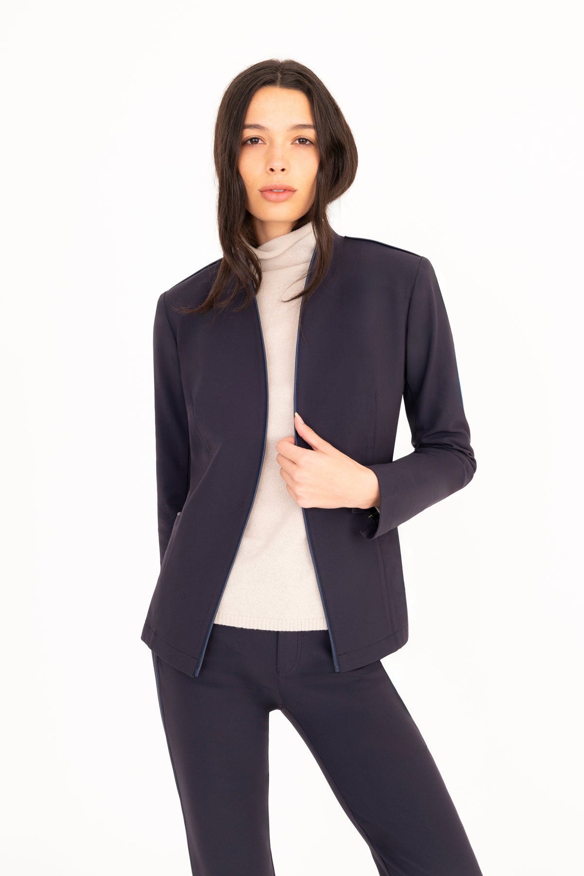 A woman modeling a dark navy blue tailored blazer and matching trousers over a cream turtleneck, photographed against a seamless white studio background.