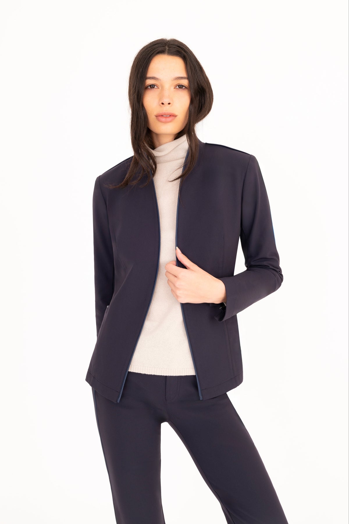 A woman modeling a dark navy blue tailored blazer and matching trousers over a cream turtleneck, photographed against a seamless white studio background.