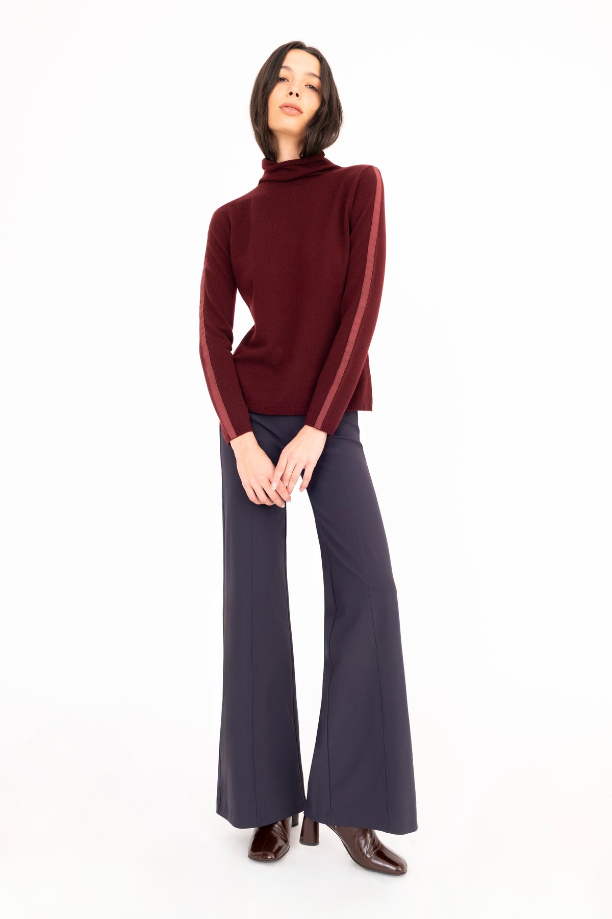 A model wearing a burgundy cashmere turtleneck, navy wide-leg trousers, and brown leather boots against a solid white background.