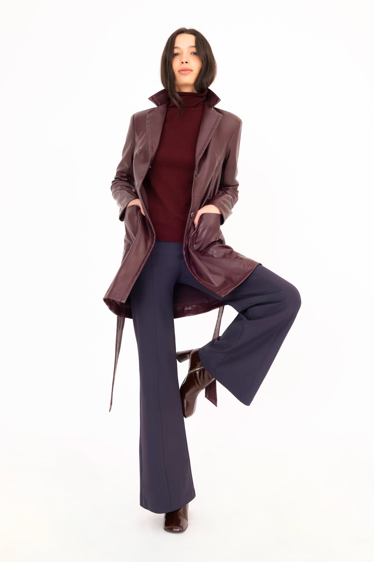 A model posing in a studio setting wearing a burgundy vegan leather trench coat over a burgundy cashmere turtleneck, navy wide-leg trousers, and brown leather boots against a solid white background.