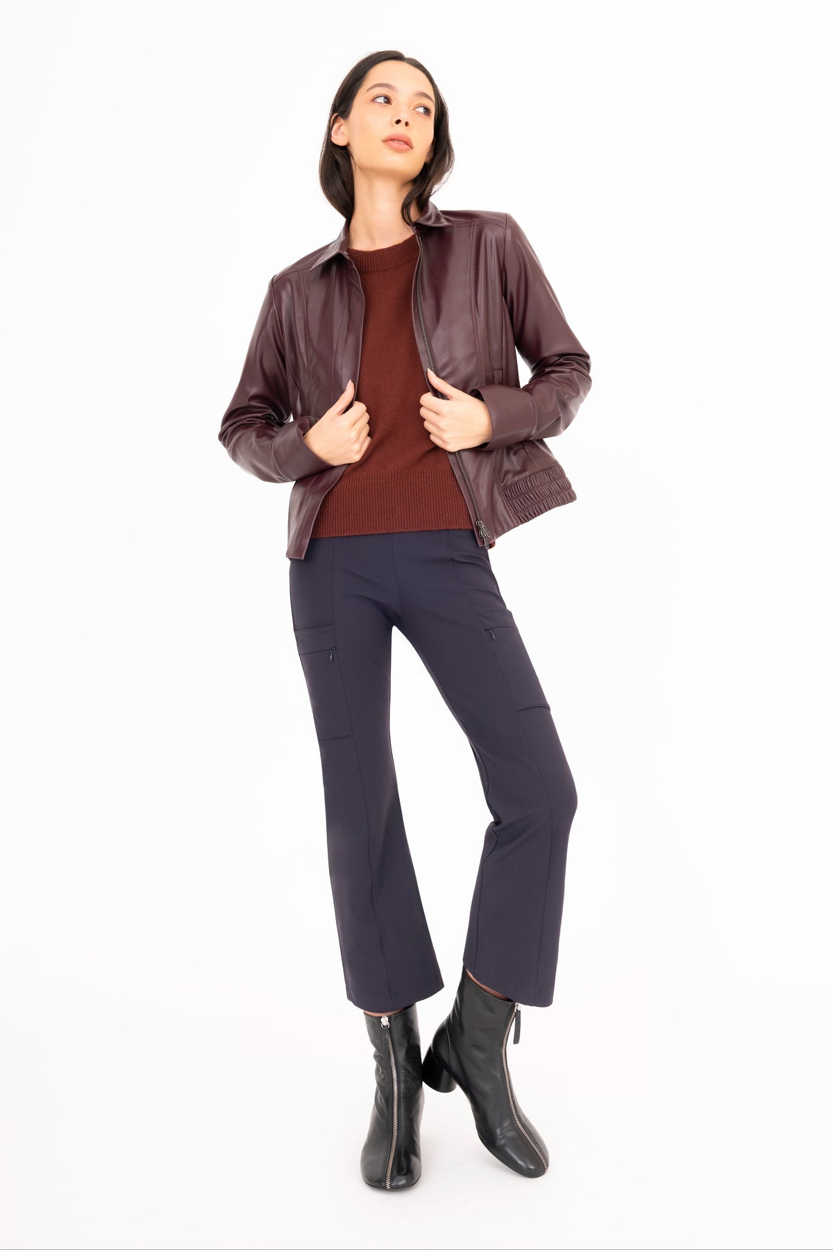 A woman modeling a dark brown leather jacket over a burgundy top, navy blue pants, and black ankle boots, standing against a solid white background.