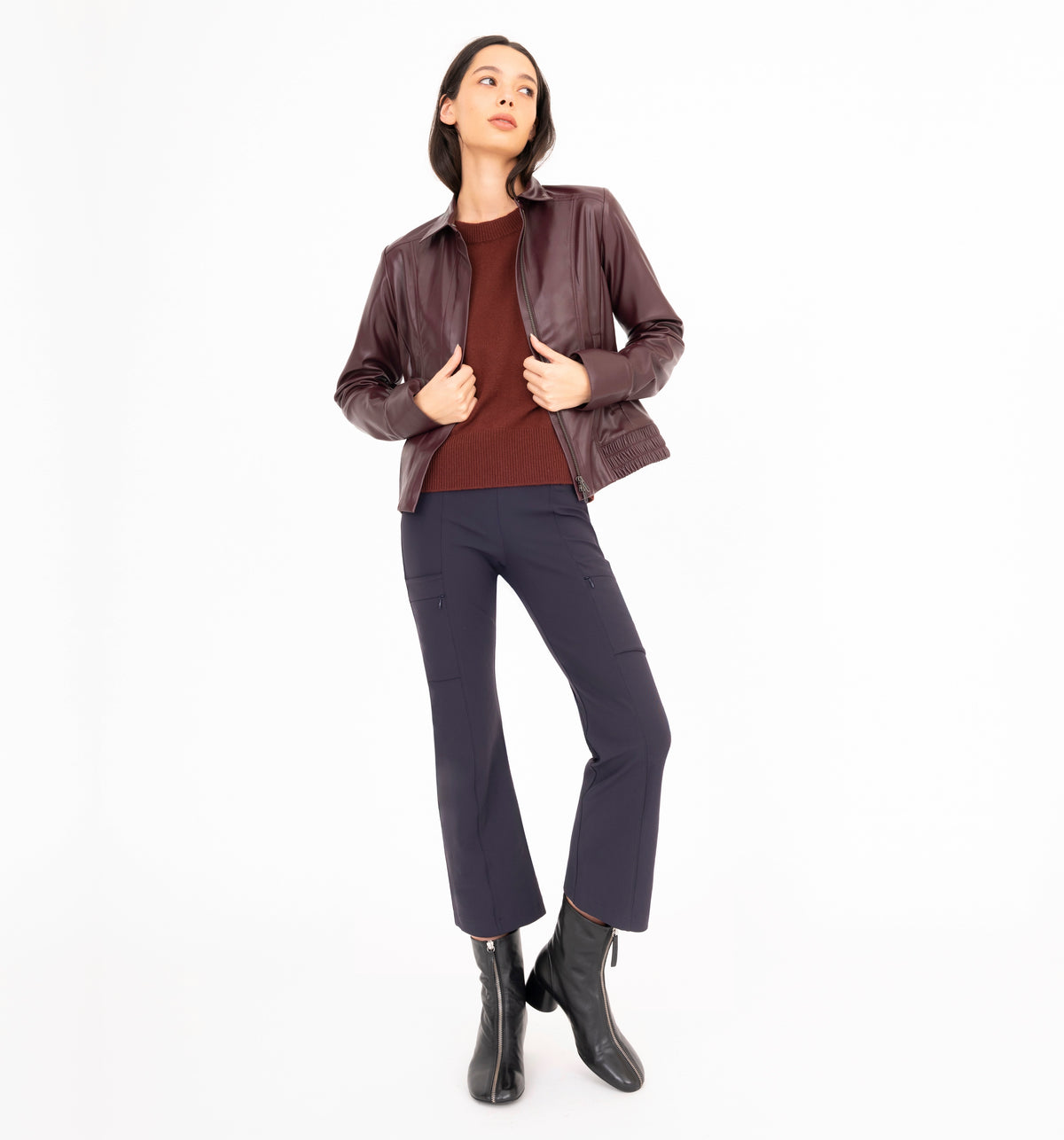 A woman modeling a dark brown leather jacket over a burgundy top, navy blue pants, and black ankle boots, standing against a solid white background.