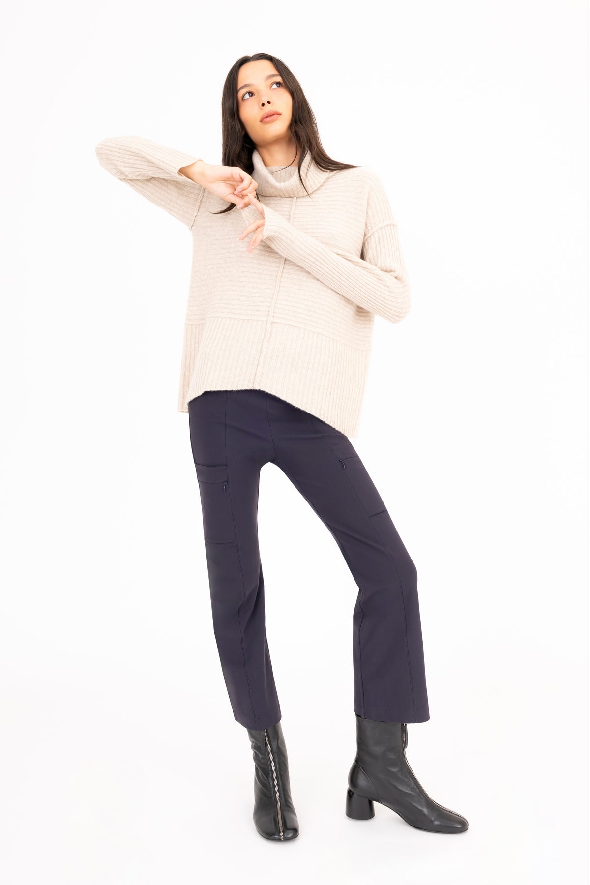 A woman modeling a cream cashmere ribbed knit turtleneck sweater, dark navy pants, and black ankle boots against a solid white background.