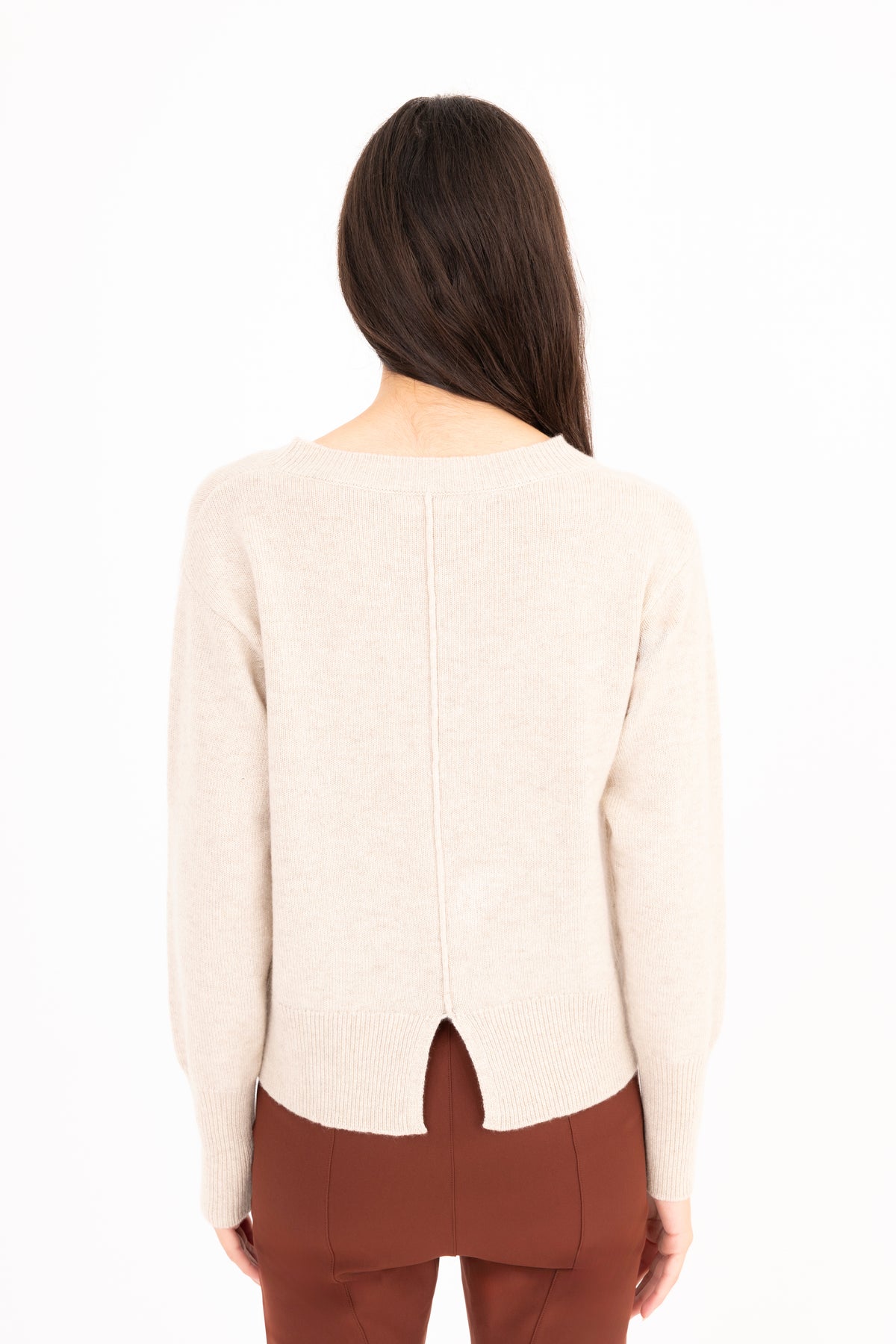 A woman viewed from the back wearing a light cream knit sweater with a split back cutout and brown pants, against a plain white studio background.
