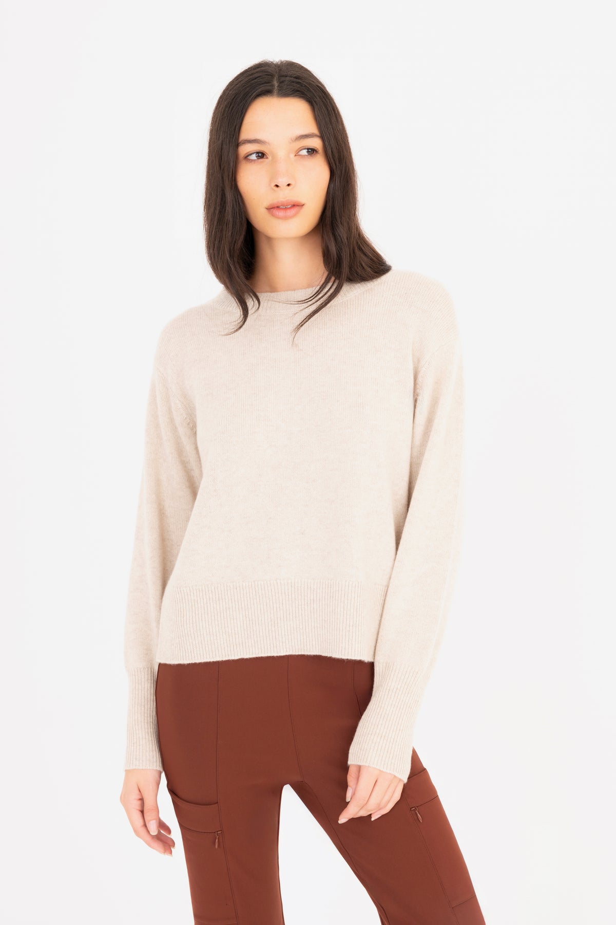 A woman modeling a light beige knit crewneck sweater paired with brown pants, shot against a seamless white studio background.