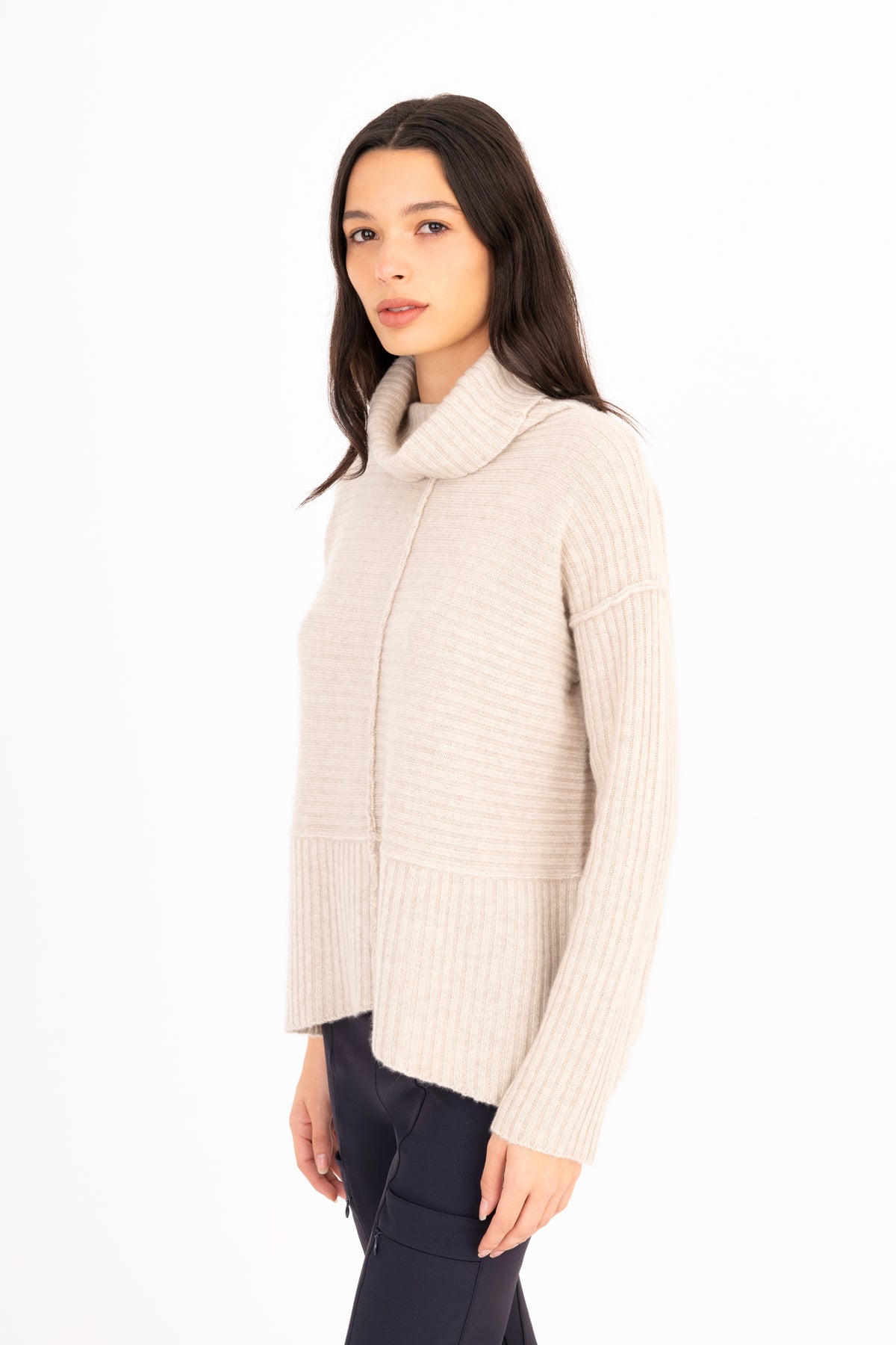 A woman modeling a ribbed knit funnel-neck sweater in a oatmeal color, paired with dark navy pants, against a plain white studio background.
