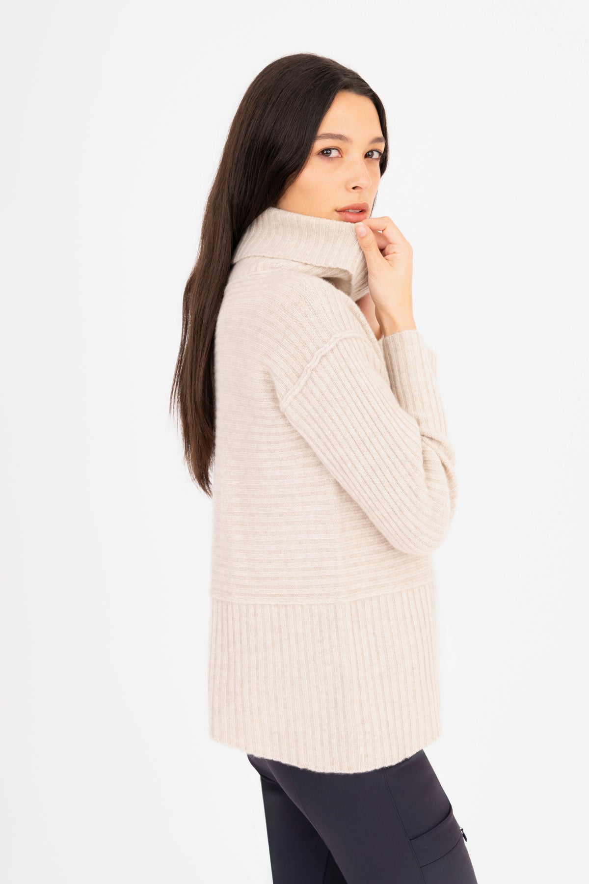 A woman modeling a ribbed knit funnel-neck sweater in a oatmeal color, paired with dark navy pants, against a plain white studio background.