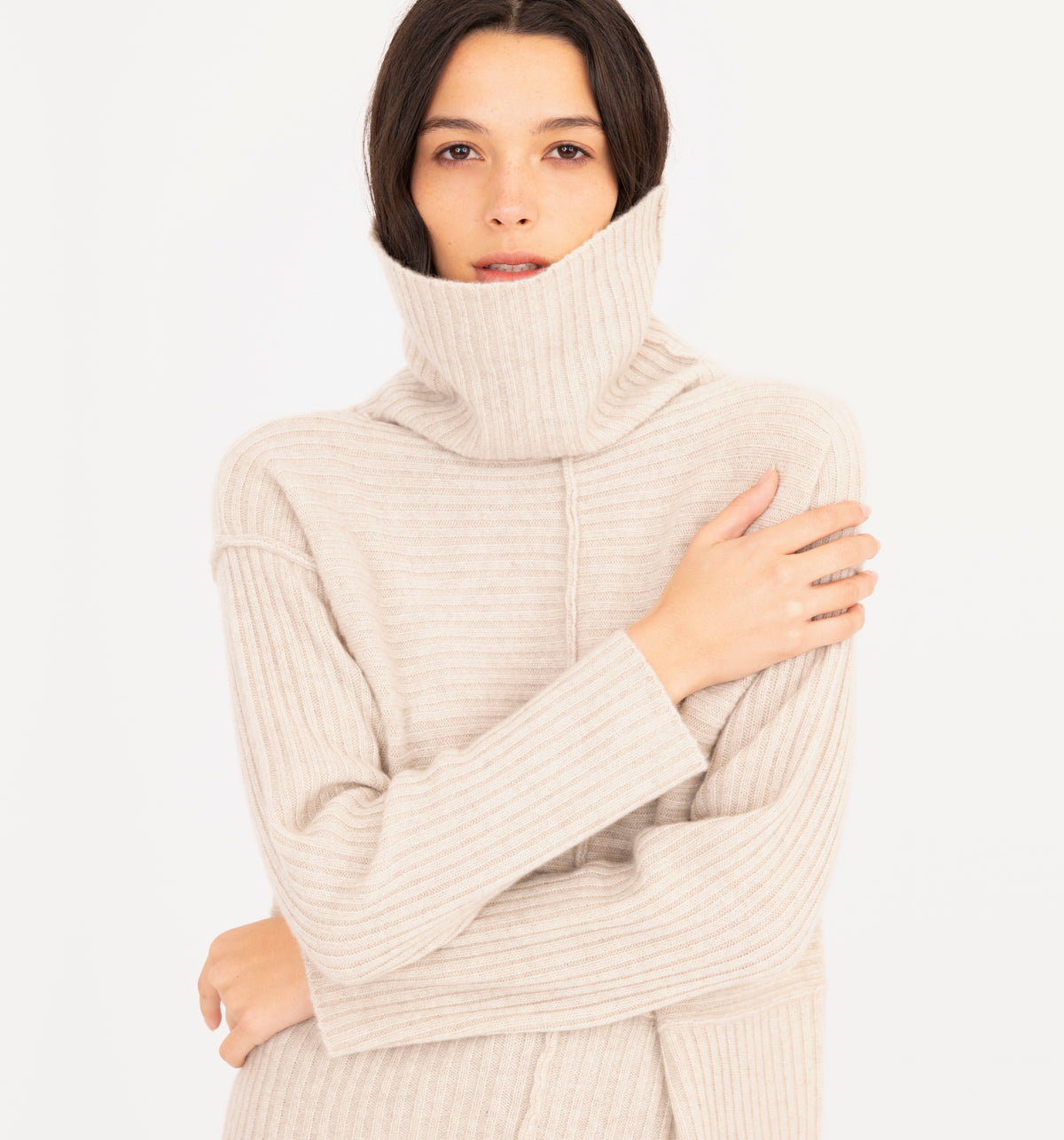 A woman wearing a cream-colored ribbed knit turtleneck sweater, posing with her arms crossed over her chest against a plain white background.