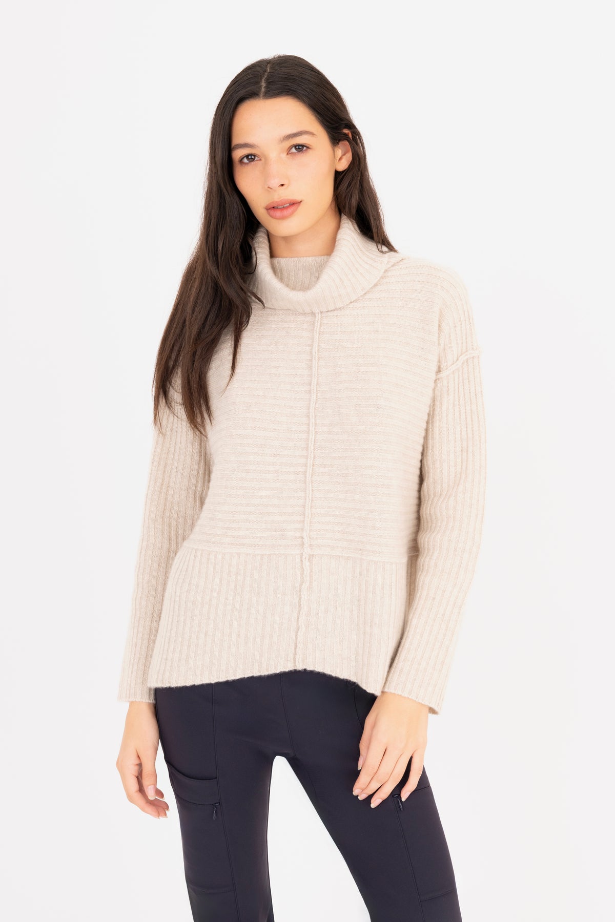 A woman modeling a ribbed knit funnel-neck sweater in a oatmeal color, paired with dark navy pants, against a plain white studio background.