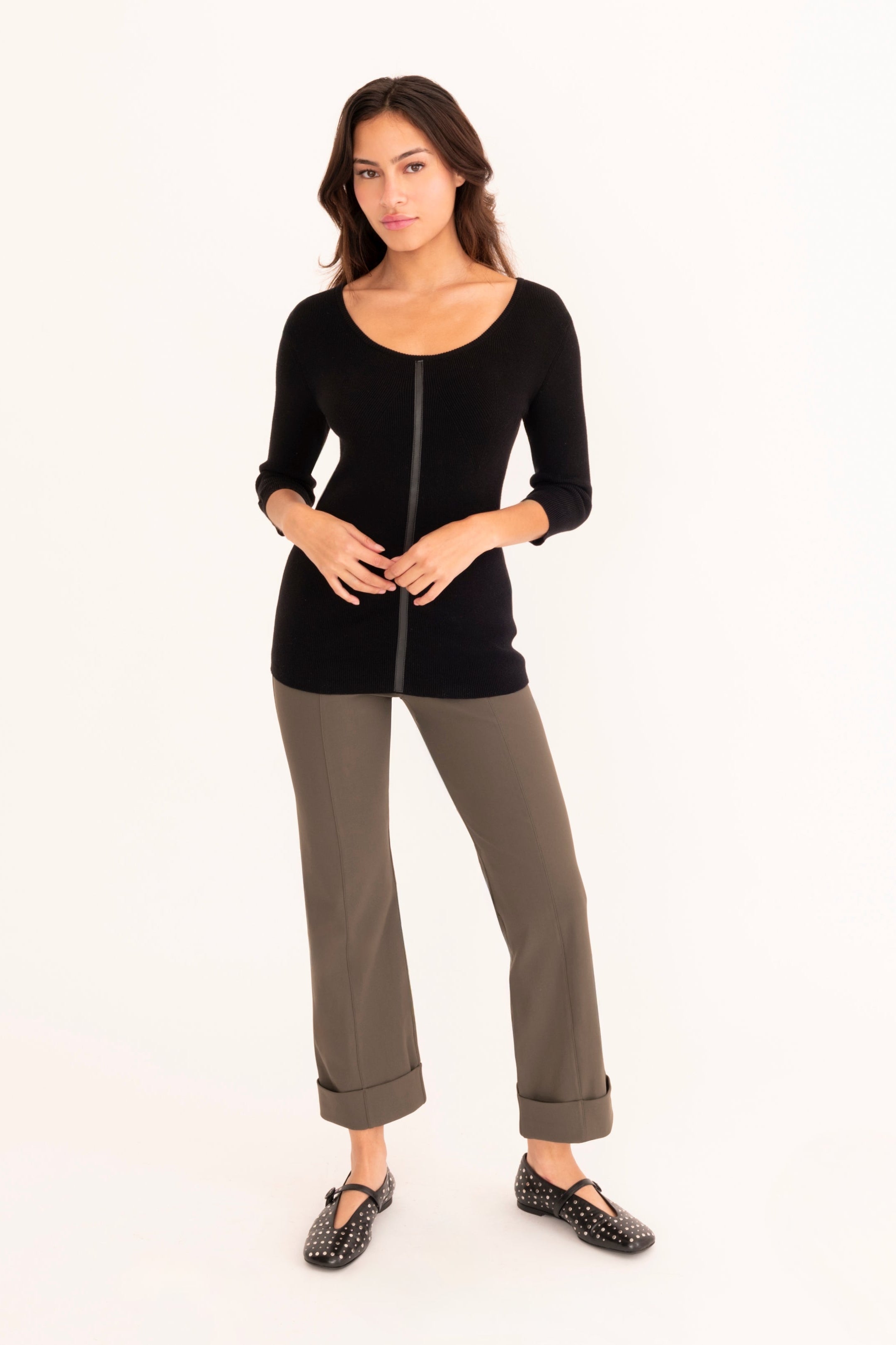 A woman modeling a black three-quarter sleeve top paired with olive green pants and black shoes, standing against a solid white background.