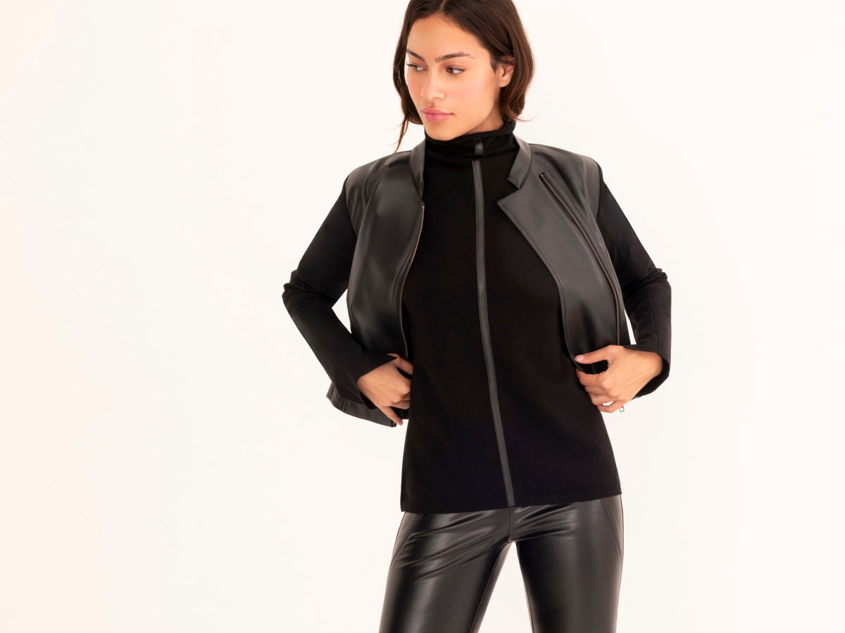 A woman modeling a black faux-leather moto jacket with a front zipper, worn over a black mock-neck top and paired with black faux-leather pants, against a plain white background.