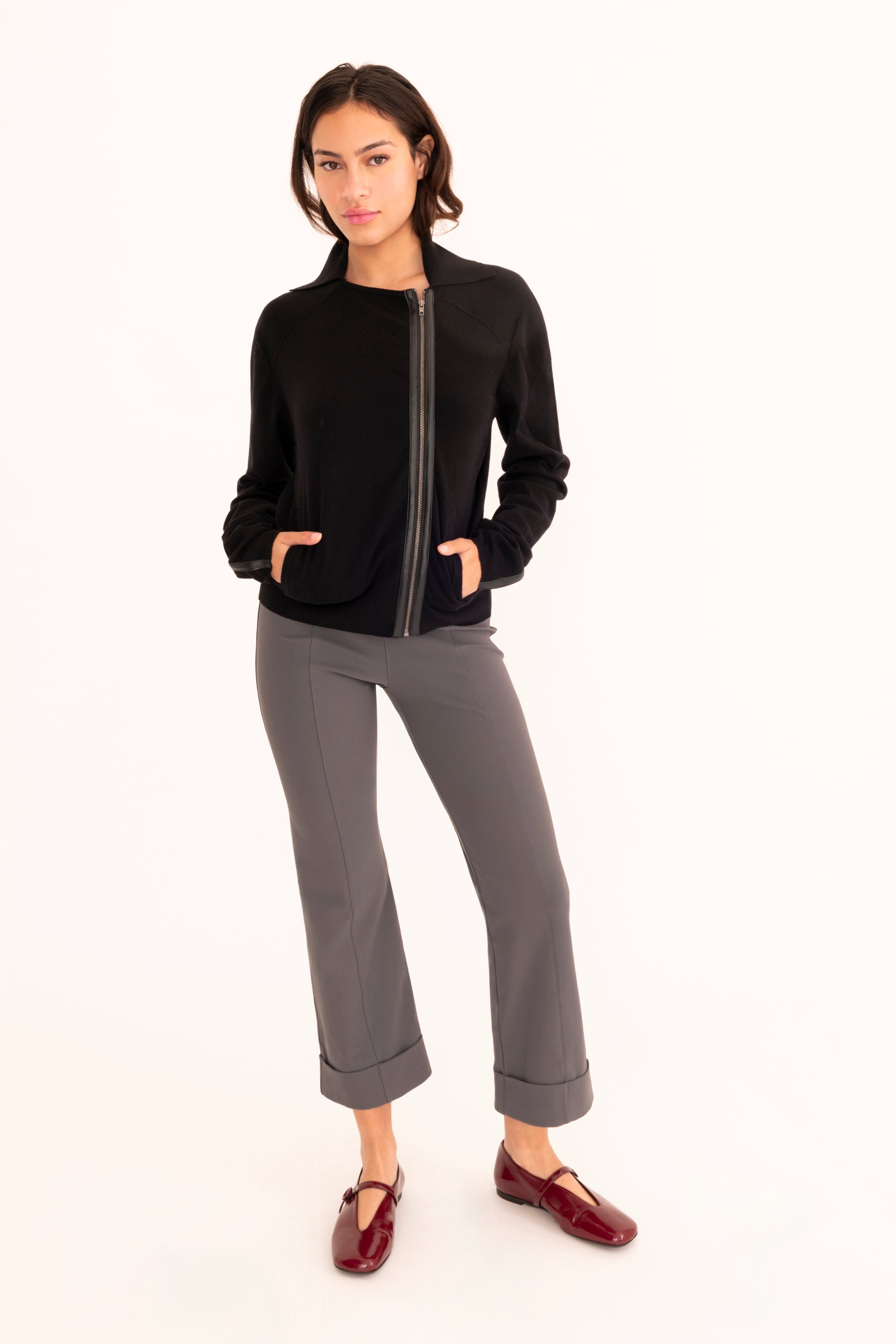 A woman modeling a black zip-up knit cardigan with gray cropped pants and burgundy loafers, standing against a plain white studio background.