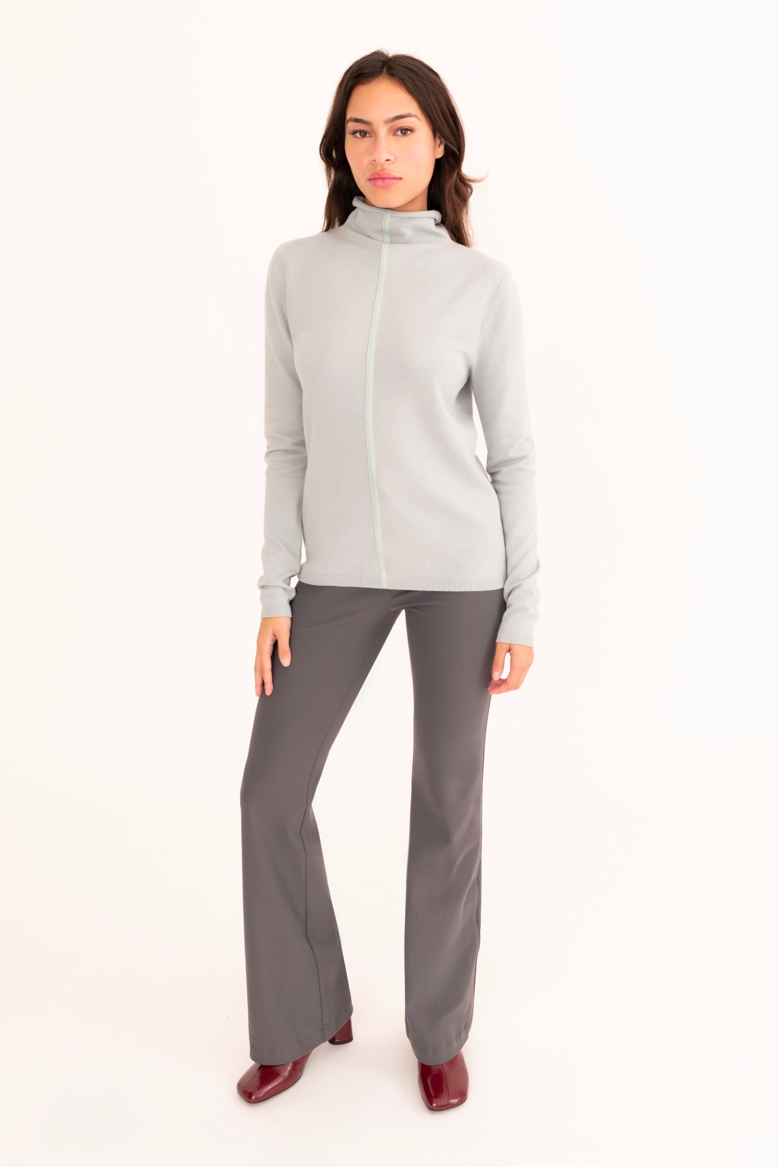 A woman modeling a light blue mock neck  sweater and dark gray pants against a plain white studio background.