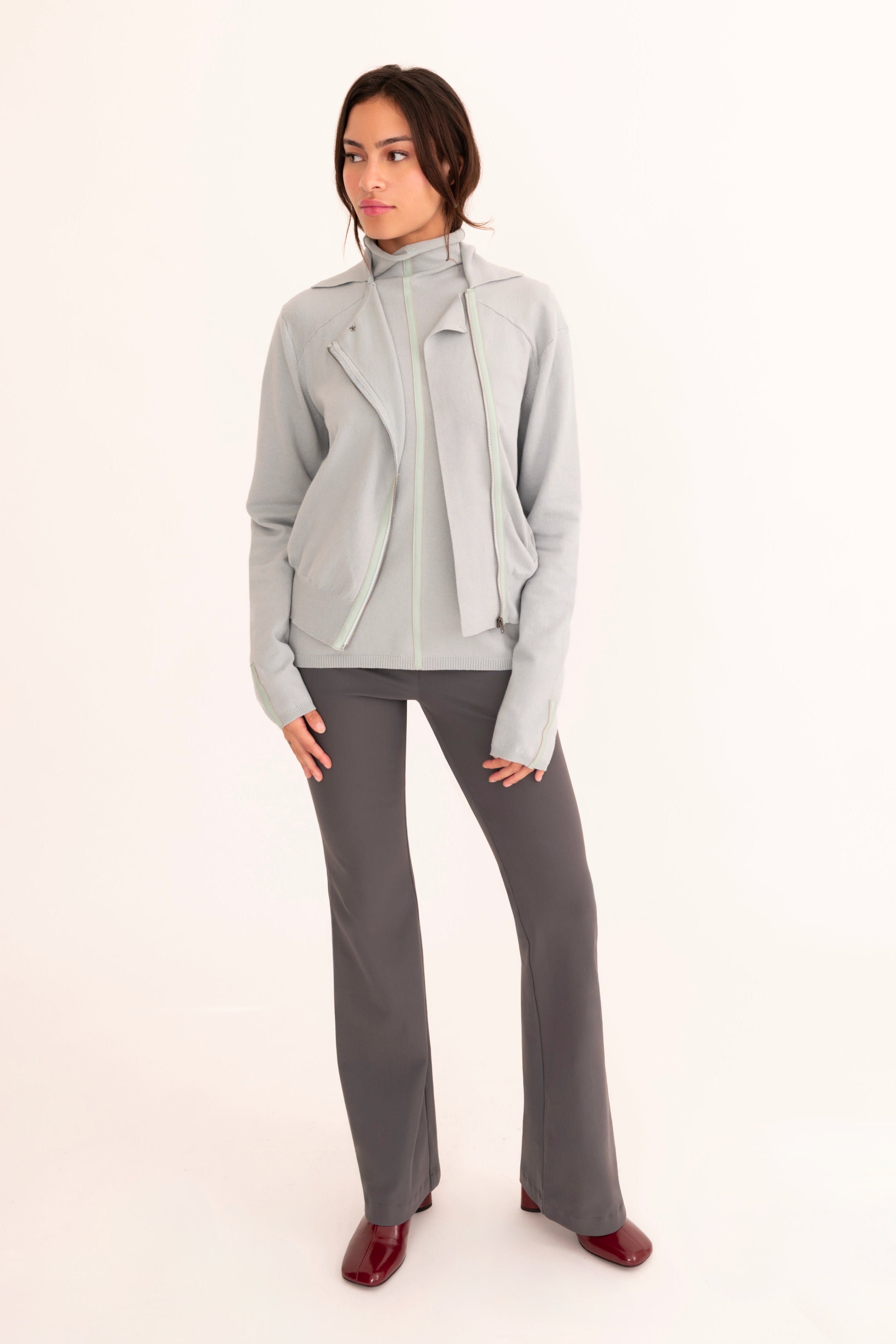 A model stands against a plain white studio background wearing a light blue jacket over a matching blue top, gray pants, and dark red shoes.